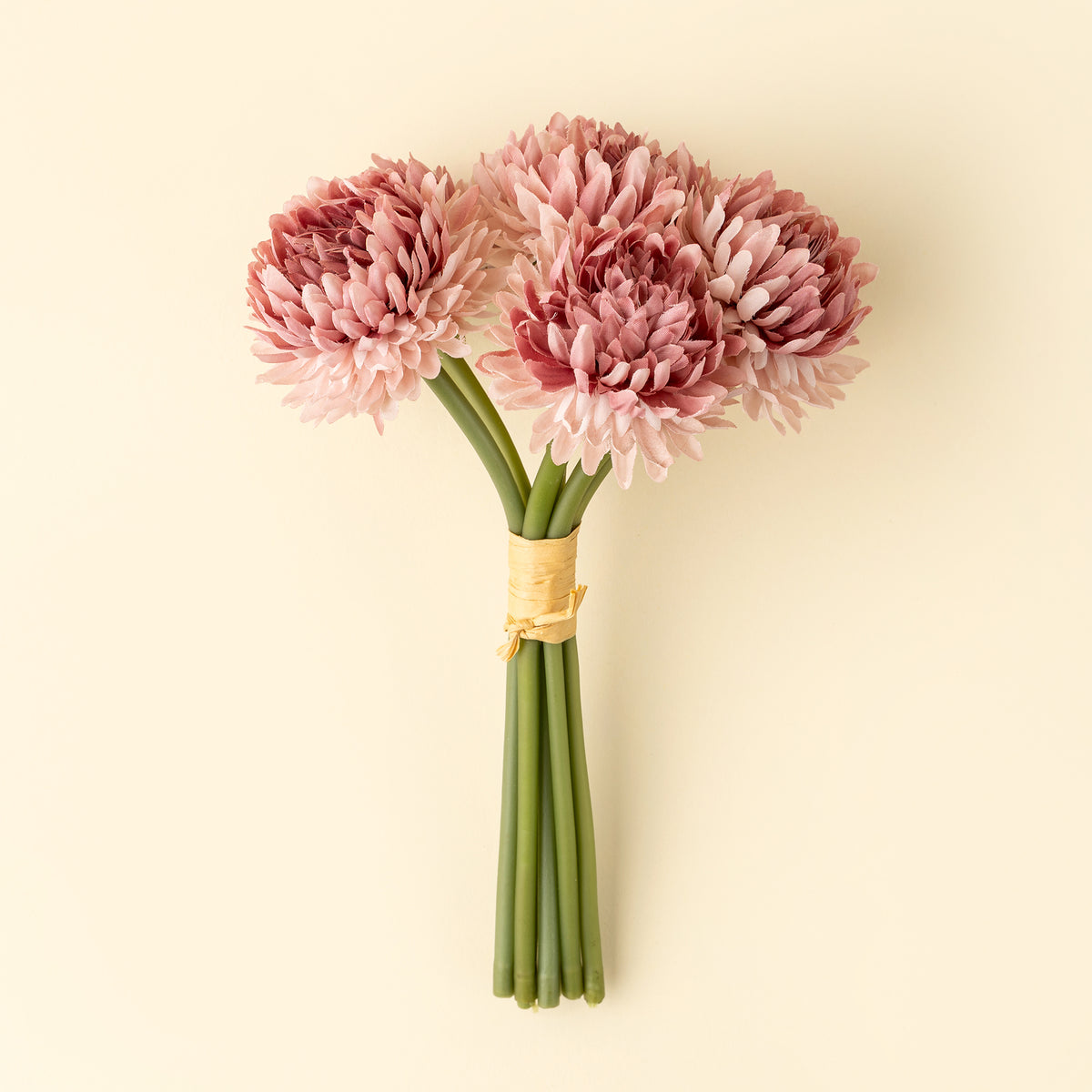 A small bouquet of pink chrysanthemum-like artificial flowers with long green stems, tied together with a natural tan band, placed on a light beige surface at la maison garçonne in montreal.