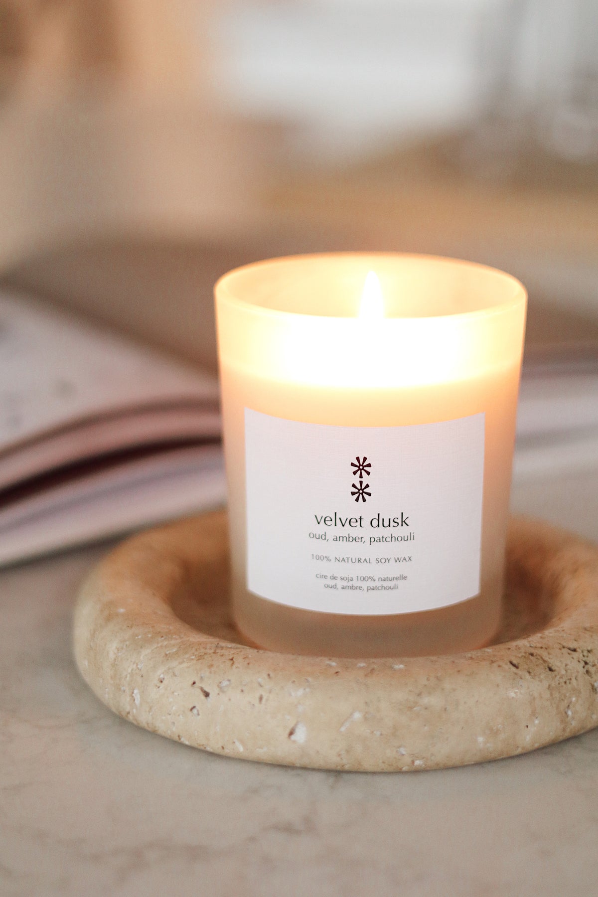 A lit candle in a clear glass jar sits on a round stone coaster. The label reads 'velvet dusk' with a small snowflake-like logo at la maison garçonne in montreal.