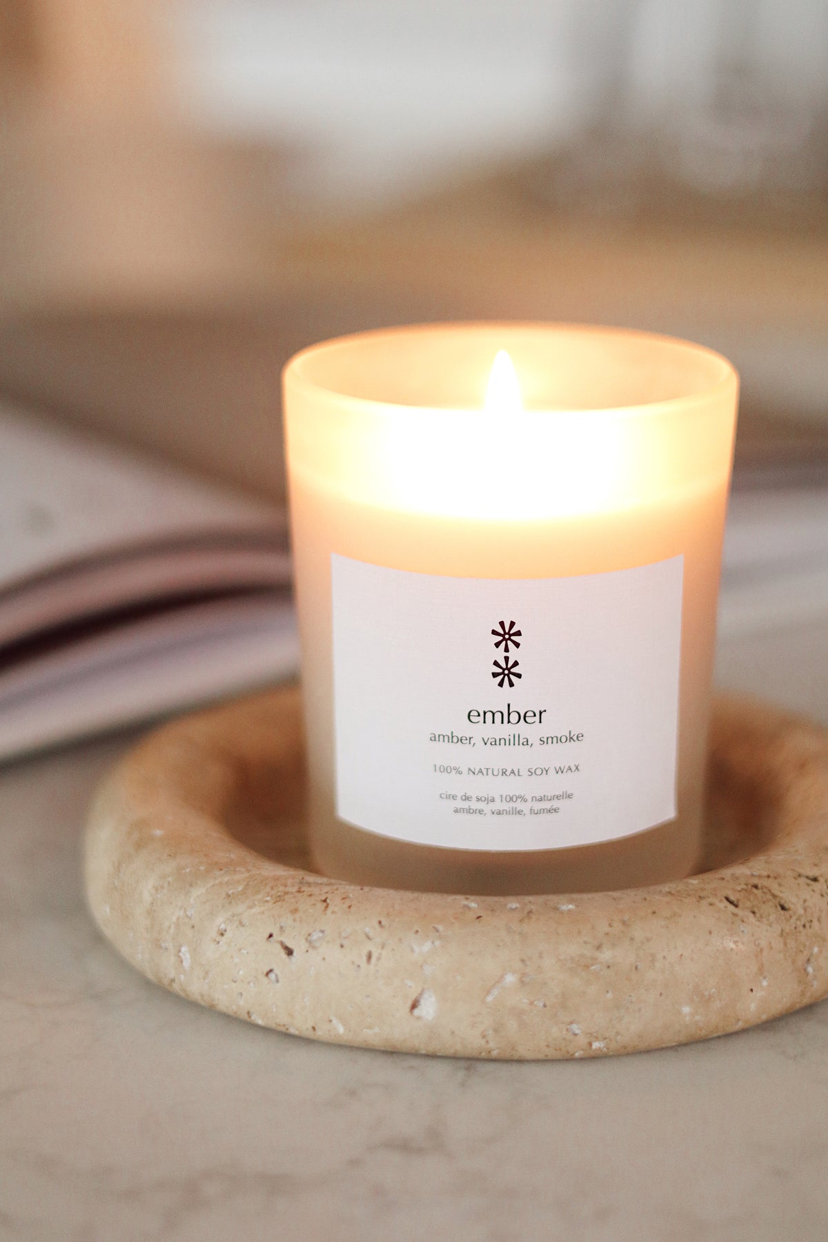A lit candle in a clear glass jar with a white label reading 'ember amber, vanilla, smoke' sits on a round beige stone coaster. The background is softly blurred with warm neutral tones at la maison garçonne in montreal.