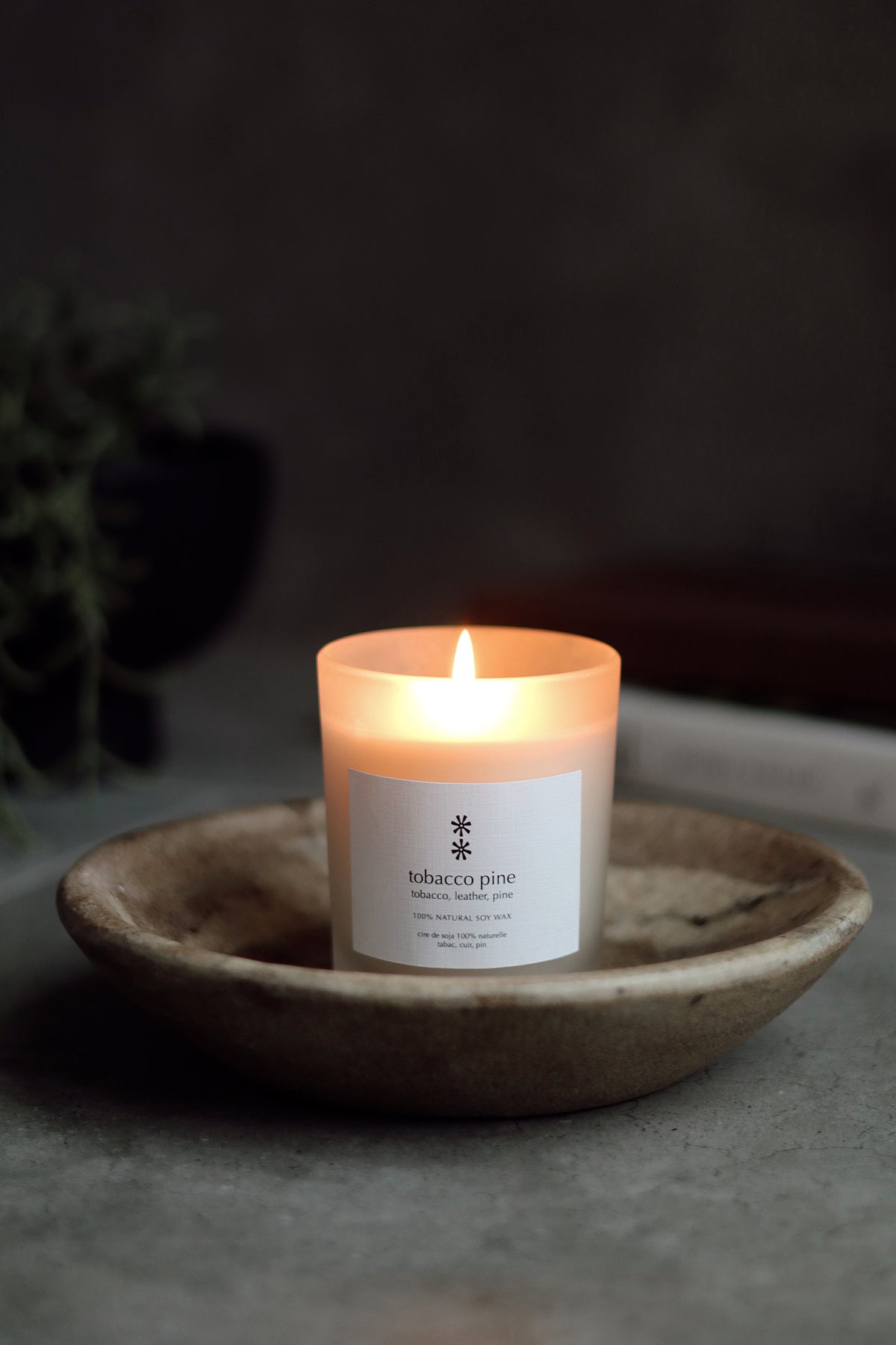 A lit candle in a white glass jar sits in a shallow round ceramic dish. The label reads 'tobacco pine' with a small snowflake-like logo at la maison garçonne in montreal.