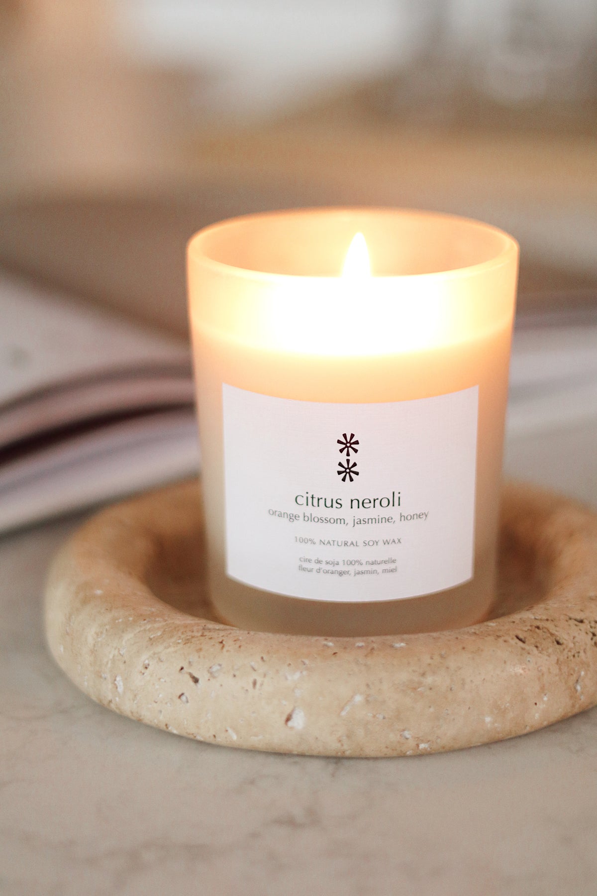 A lit candle in a frosted glass jar with a white label reading 'citrus neroli' and 'orange blossom, jasmine, honey,' resting on a round stone coaster at la maison garçonne in montreal