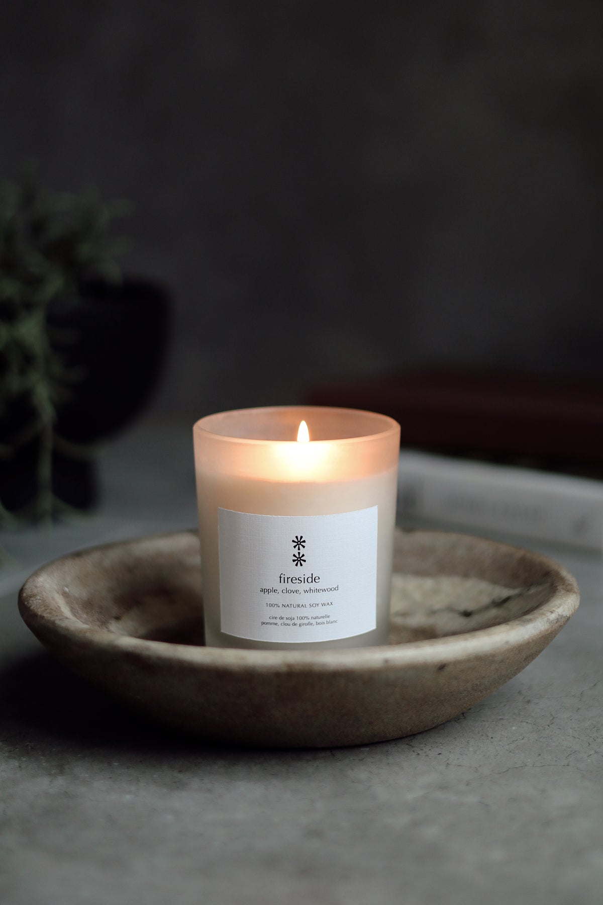A lit candle in a frosted glass jar sits in a shallow ceramic dish on a gray stone surface. The label reads 'fireside' with a small snowflake-like symbol above it at la maison garçonne.