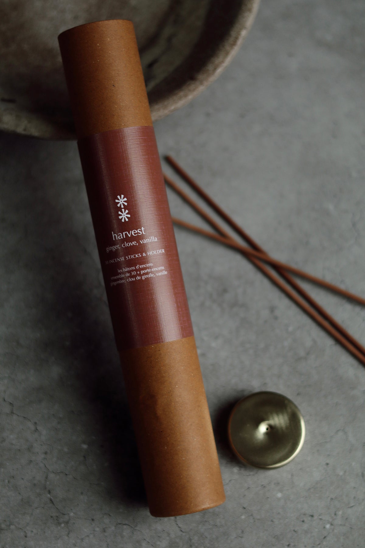 A cylindrical brown kraft-paper tube with a dark brown label reading 'harvest' and 'ginger, jasmine, sandalwood incense sticks' lies on a gray surface. A small round metal cap and several incense sticks are placed nearby, with a blurred ceramic bowl in the background at la maison garçonne in montreal.