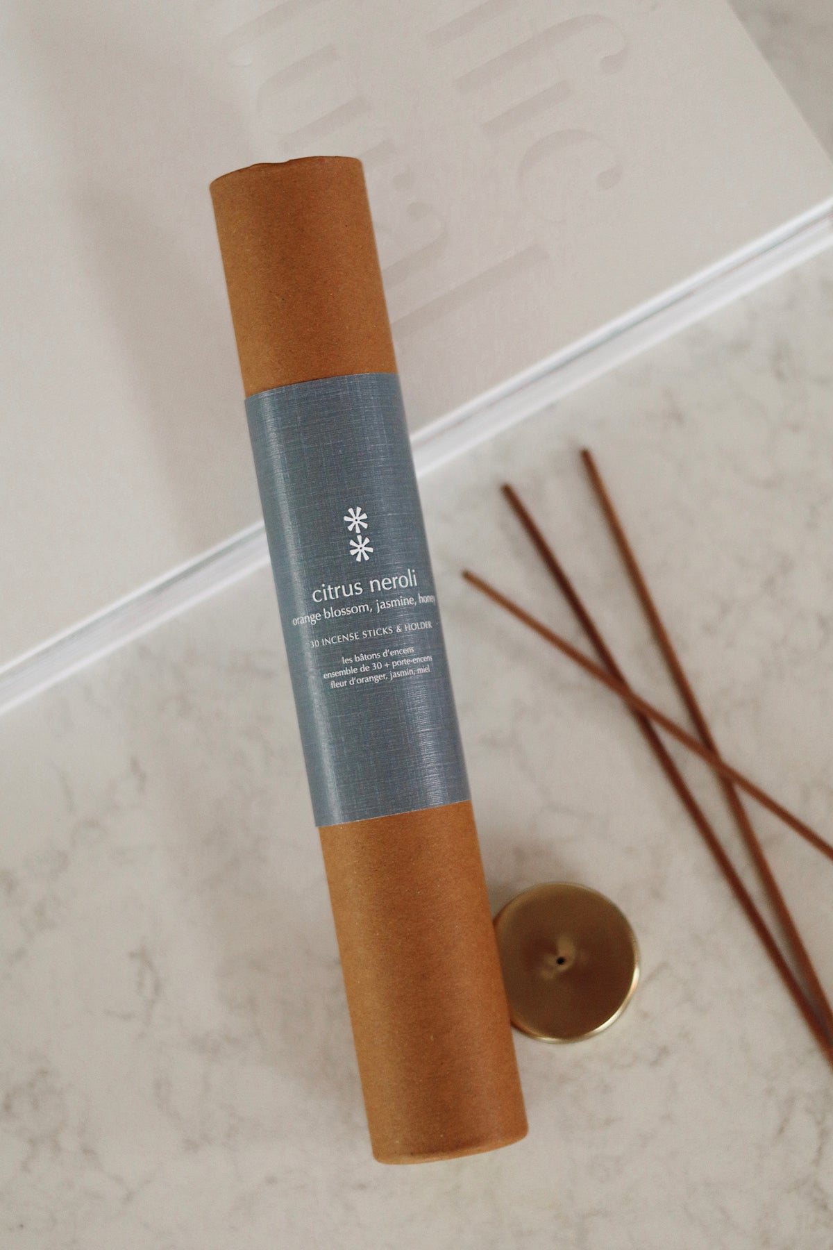 A rolled brown kraft paper tube labeled 'citrus neroli' with a gray paper band, placed on a light marble surface beside three wooden diffuser sticks and a small gold cap at la maison garçonne in montreal.