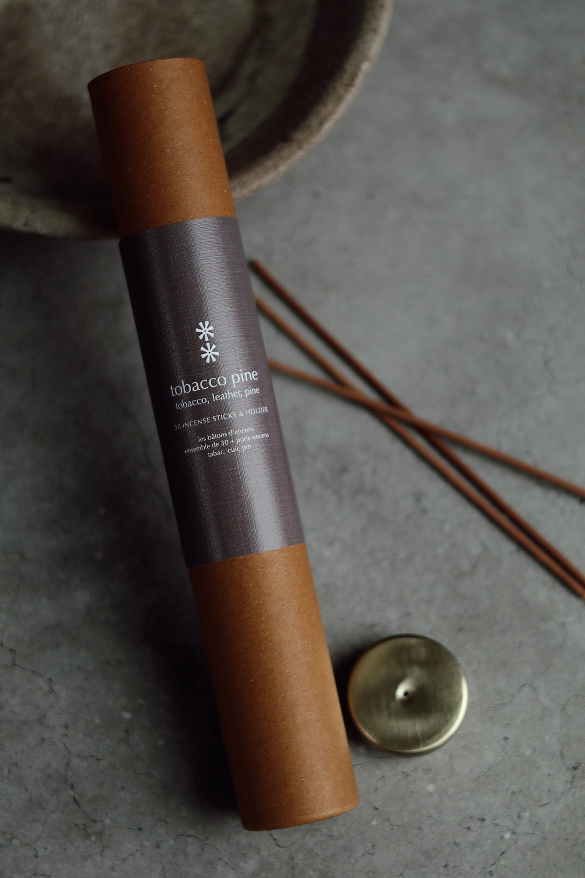 A roll of incense sticks in a brown kraft paper tube with a dark label reading 'tobacco pine', placed on a gray surface with a small brass incense burner and incense sticks nearby at la maison garçonne in montreal.