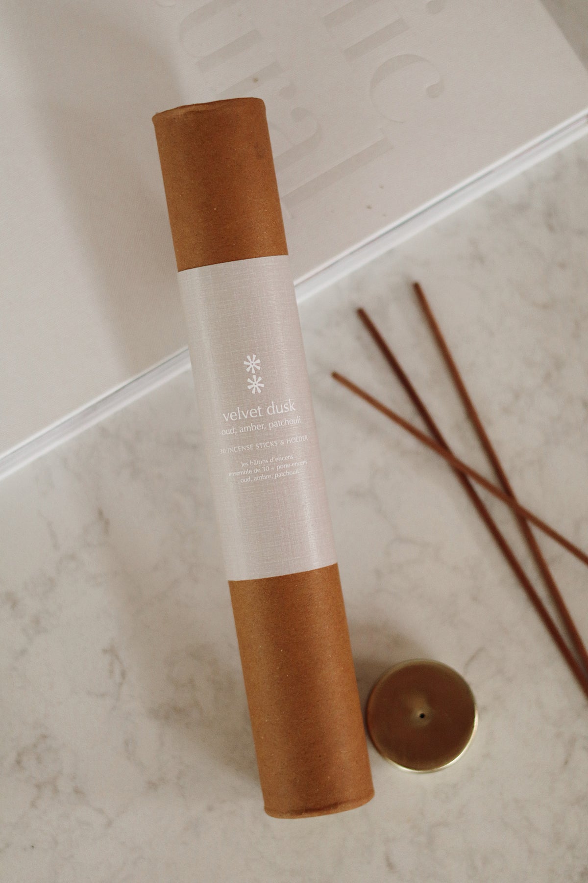 A rolled brown candle in a kraft paper sleeve with a white label, placed on a light marble surface next to two wooden diffuser sticks and a small round metal tin at la maison garçonne in montreal.