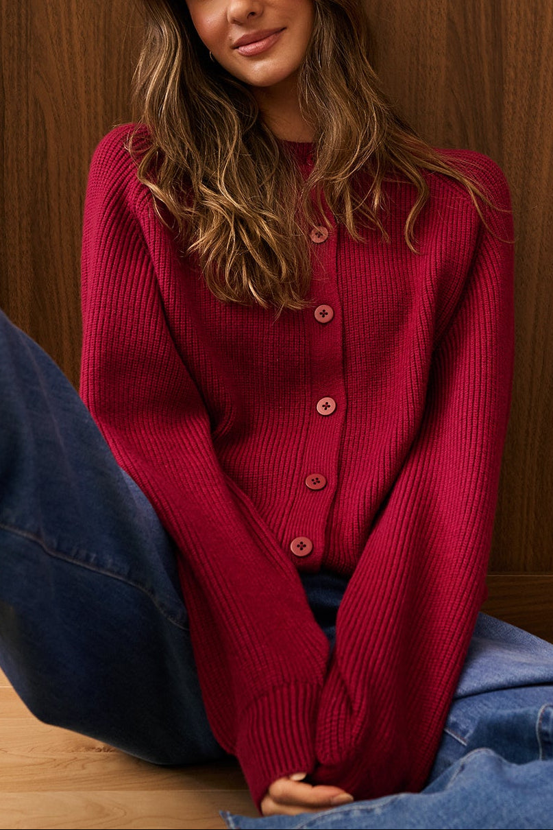 A woman sits on a light wood floor in front of a dark wood-paneled wall, wearing Alayna Burgundy Oversized Knit Cardigan by Dailystory available at la petite garçonne