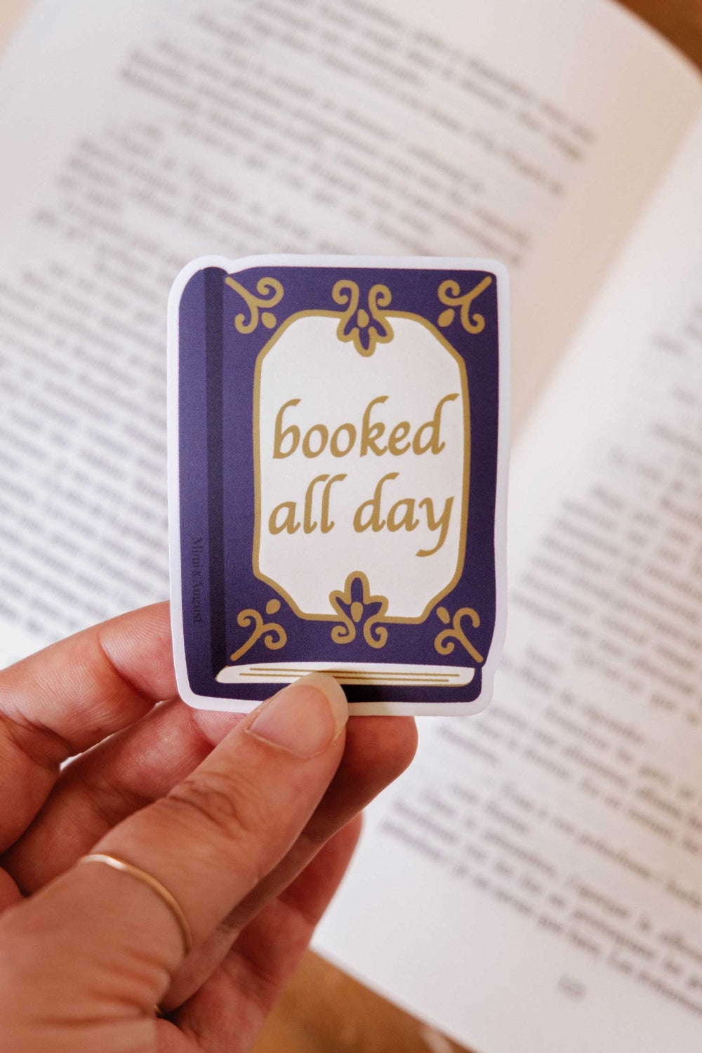 woman holding a Small book sticker with 'booked all day' from mimi and august on a page of an open book