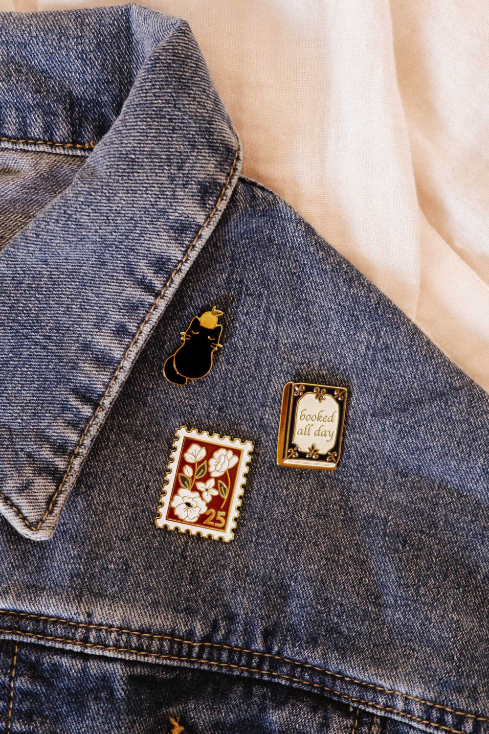 Denim jacket with decorative Enamel pins from Mimi and August featuring a vintage stamp design and a book all day.