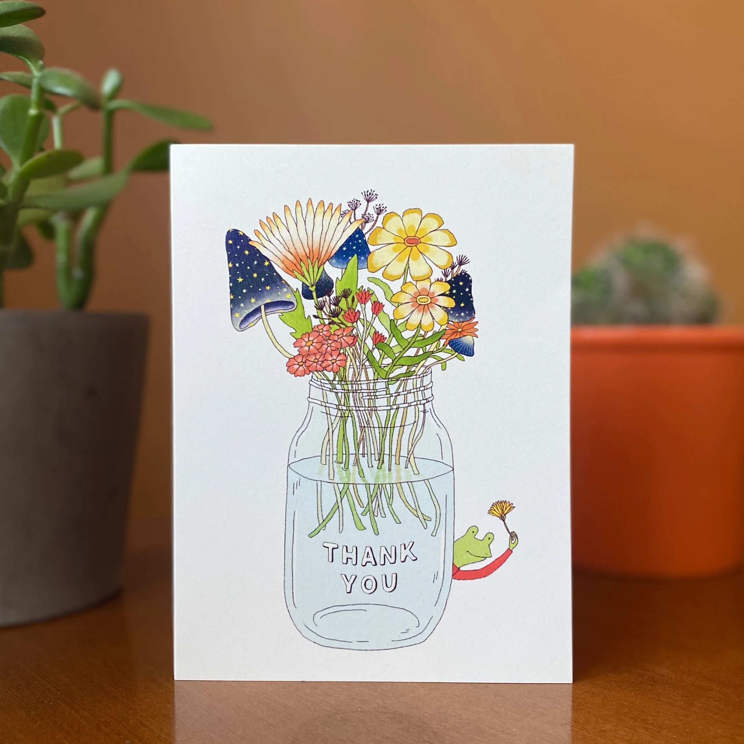 Greeting card with a mason jar of flowers and 'Thank You' text, placed on a wooden surface with plants in the background.