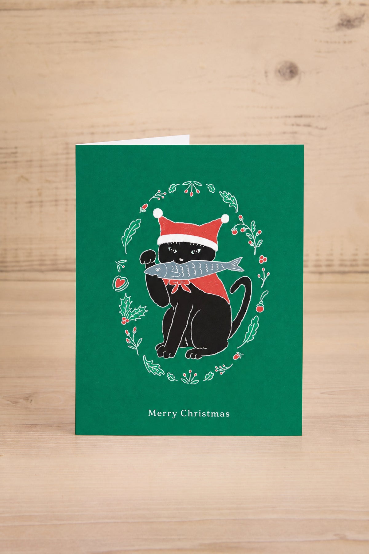 A green Christmas greeting card by Foonie featuring a black cat wearing a Santa hat and holding a fish, surrounded by a holly wreath. The card is displayed standing upright on a light-colored wooden surface at la maison garçonne in montreal.
