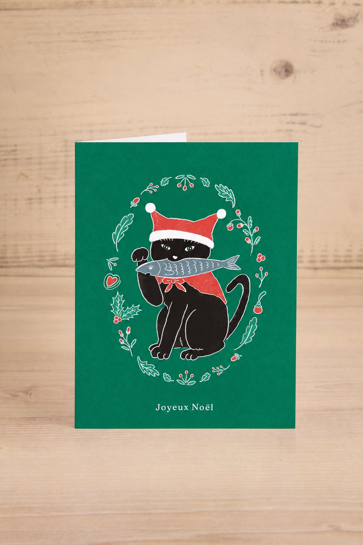 A green Christmas french greeting card by Foonie featuring a black cat wearing a Santa hat and holding a fish, surrounded by a holly wreath. The card is displayed standing upright on a light-colored wooden surface at la maison garçonne in montreal.
