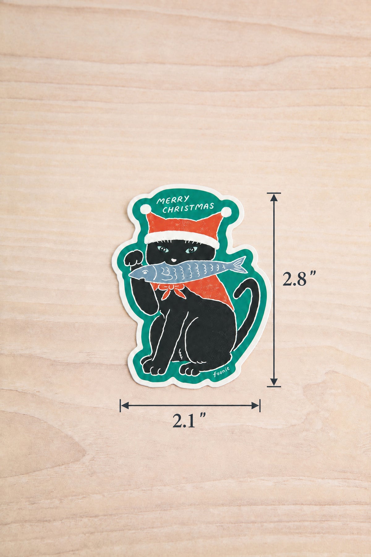 A Christmas-themed die-cut sticker from Foonie of a black cat wearing a red Santa hat and holding a silver fish, displayed on a light wood background with dimension lines indicating 2.1 inches by 2.