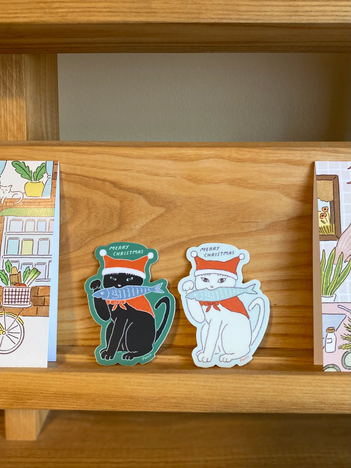 Two Christmas-themed die-cut stickers from Foonie displayed on a wooden shelf. One sticker features a black cat wearing a green hat and scarf with the text 'MERRY CHRISTMAS'.