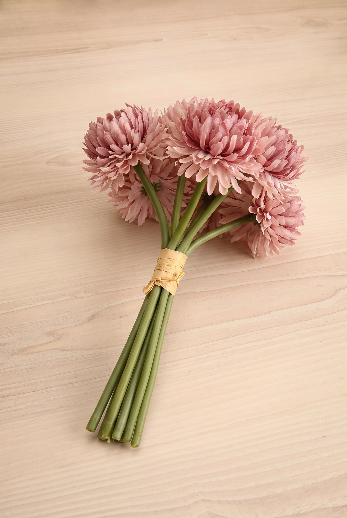 A small bouquet of pink chrysanthemum-like artificial flowers with long green stems, tied together with a natural tan band, placed on a light wood surface at la maison garçonne in montreal.
