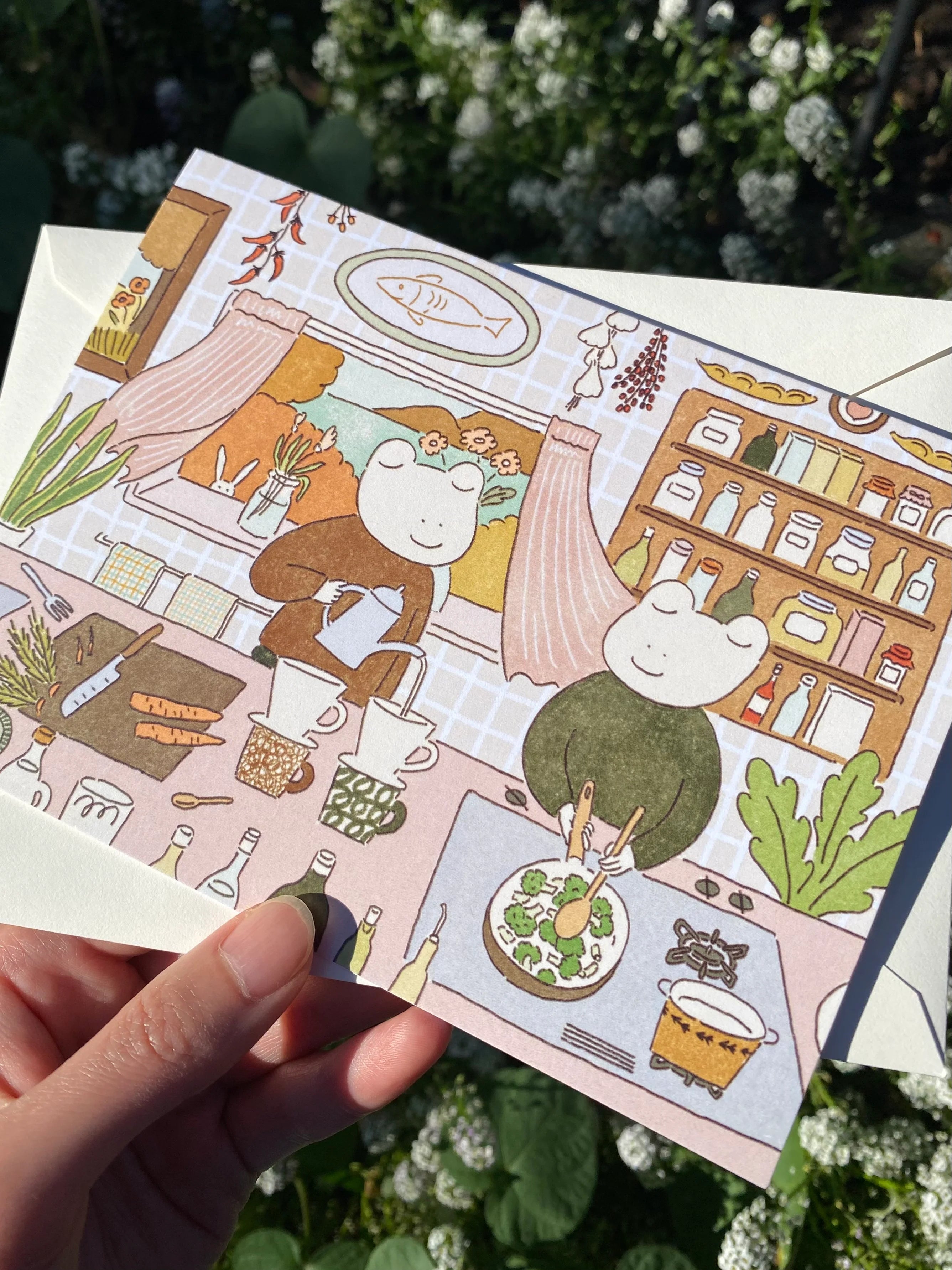 Illustrated postcard with 2 frogs in a kitchen setting, held by a hand with a blurred green background.