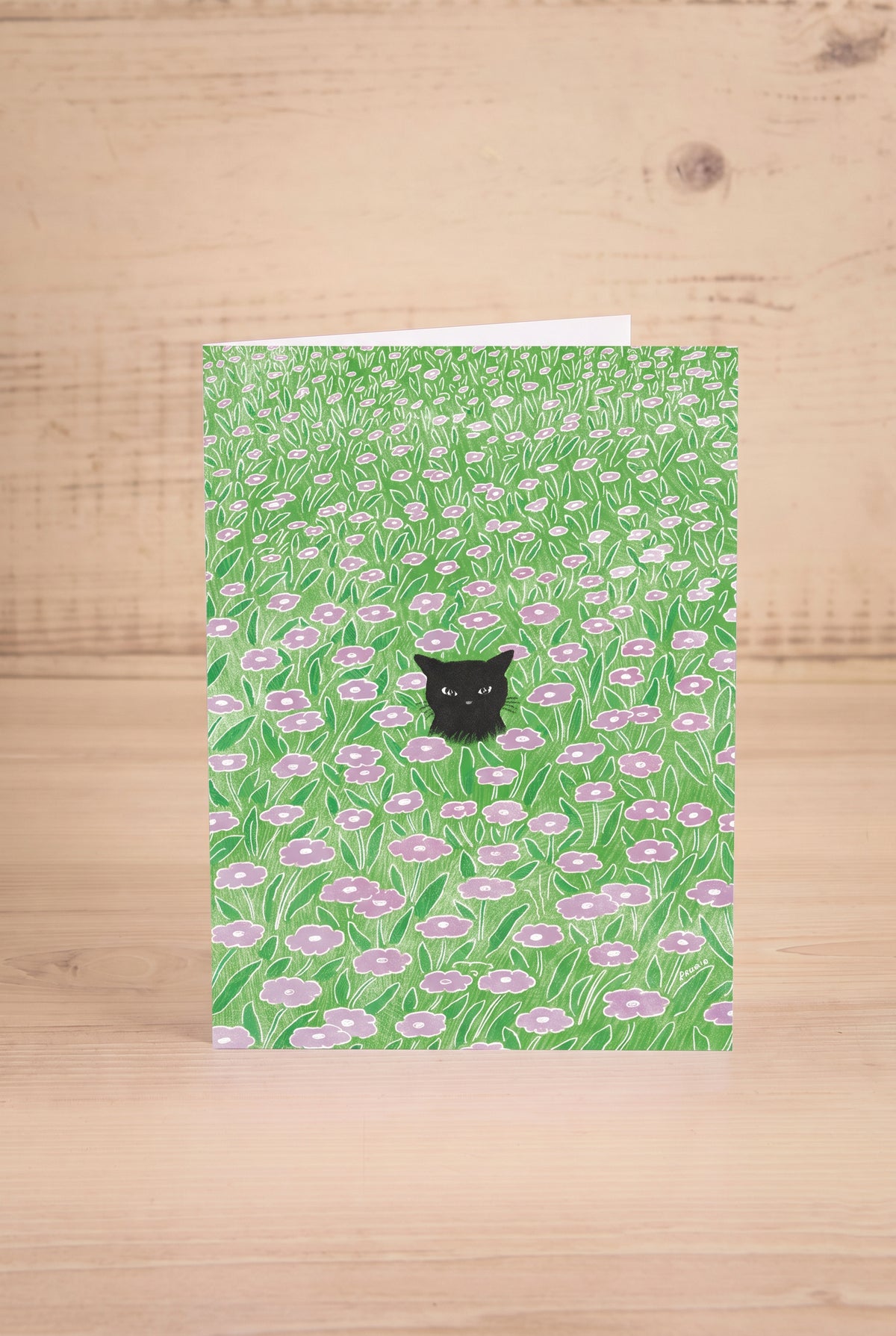 A vertical greeting card featuring a watercolor-style green field of flowers with a small black cat by Foonie at maison garçonne in montreal