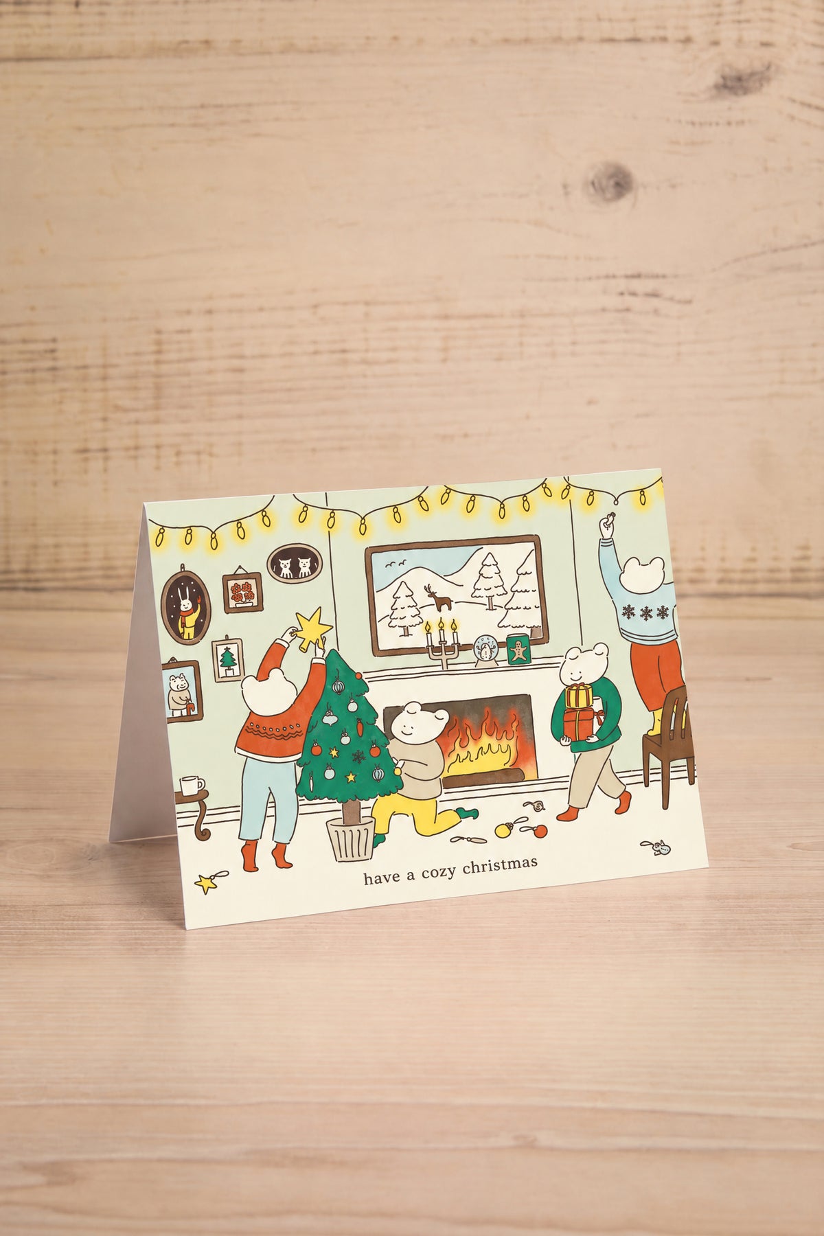 A folded Christmas greeting card by foonie with a colorful cartoon illustration of people by a fireplace and a decorated tree, standing on a light wood surface at maison garçonne montreal