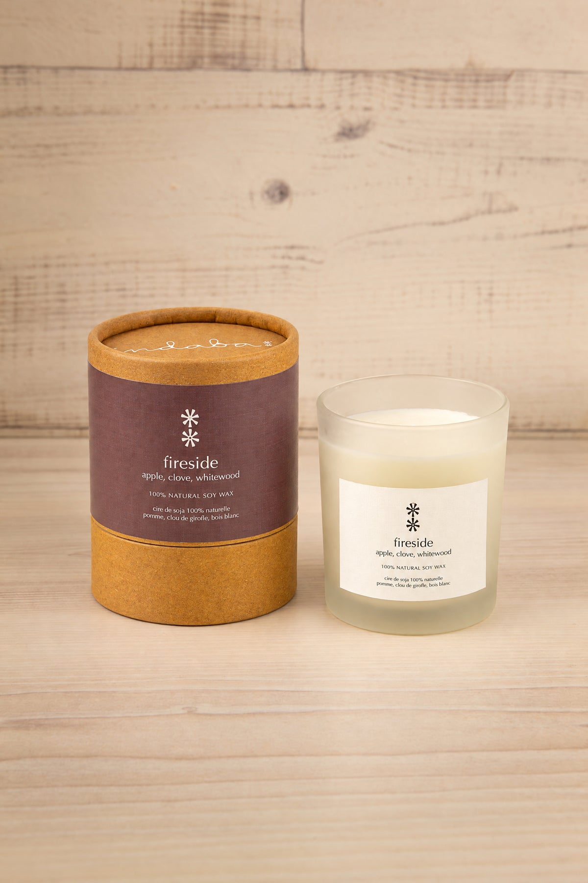 A candle in a clear glass jar next to its kraft paper packaging cylinder, both labeled 'fireside', placed on a light wood surface against a light wood plank background at la maison garçonne.