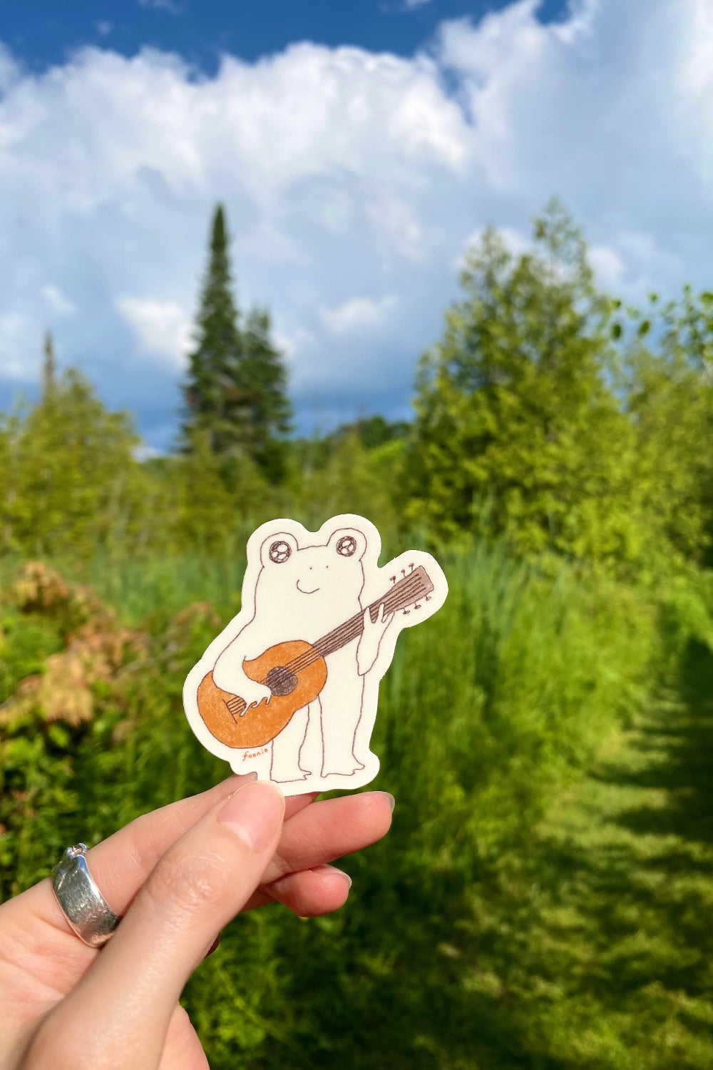 Hand holding a sticker of a frog playing a guitar against a natural background with trees and blue sky.