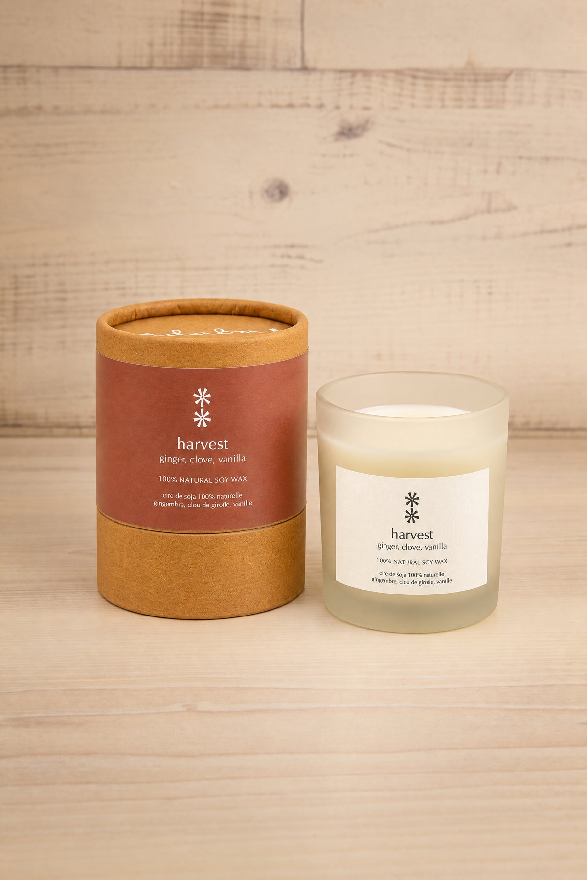 A jar candle and its cylindrical cardboard packaging container on a light wood surface against a light wood plank background. The container has a terracotta brown label with white text, and the candle jar is frosted with a white label at la maison garçonne in montreal.
