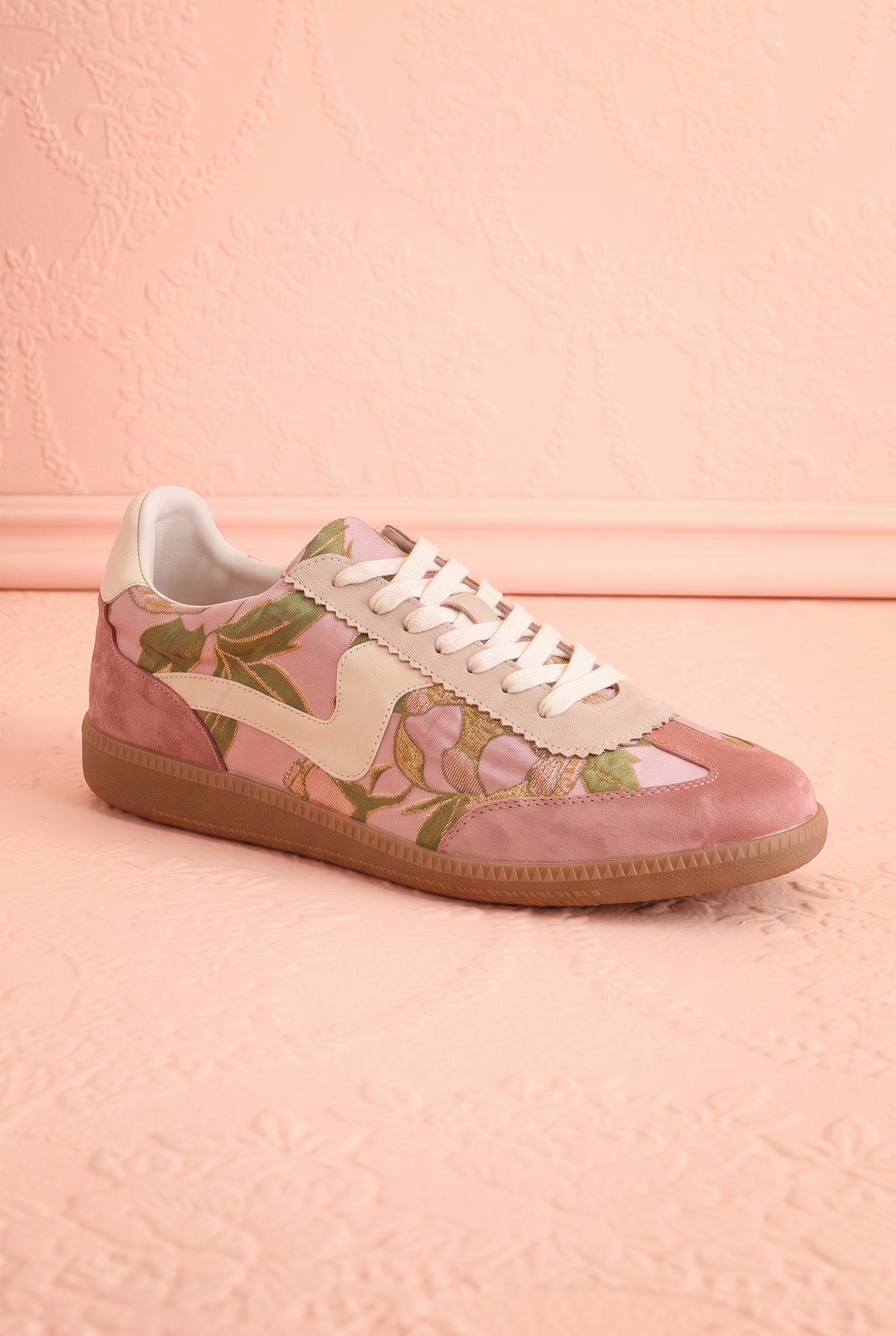 Side view of a pair of low-top sneakers in dusty pink suede and glossy pink patent leather, decorated with a green floral print, cream laces, and gum soles, displayed on a textured pink background at boutique 1861.