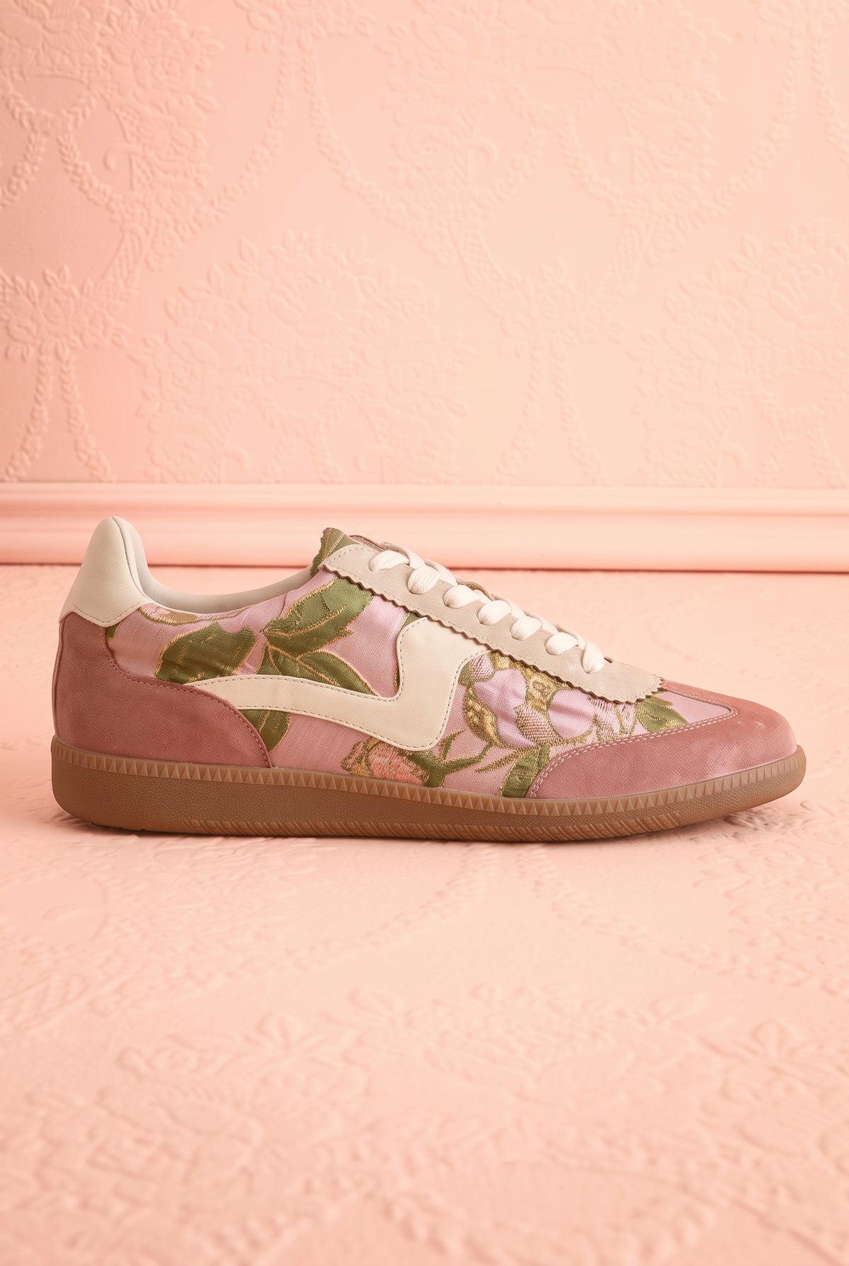 Side view of a pair of low-top sneakers in dusty pink suede and glossy pink patent leather, decorated with a green floral print, cream laces, and gum soles, displayed on a textured pink background at boutique 1861.
