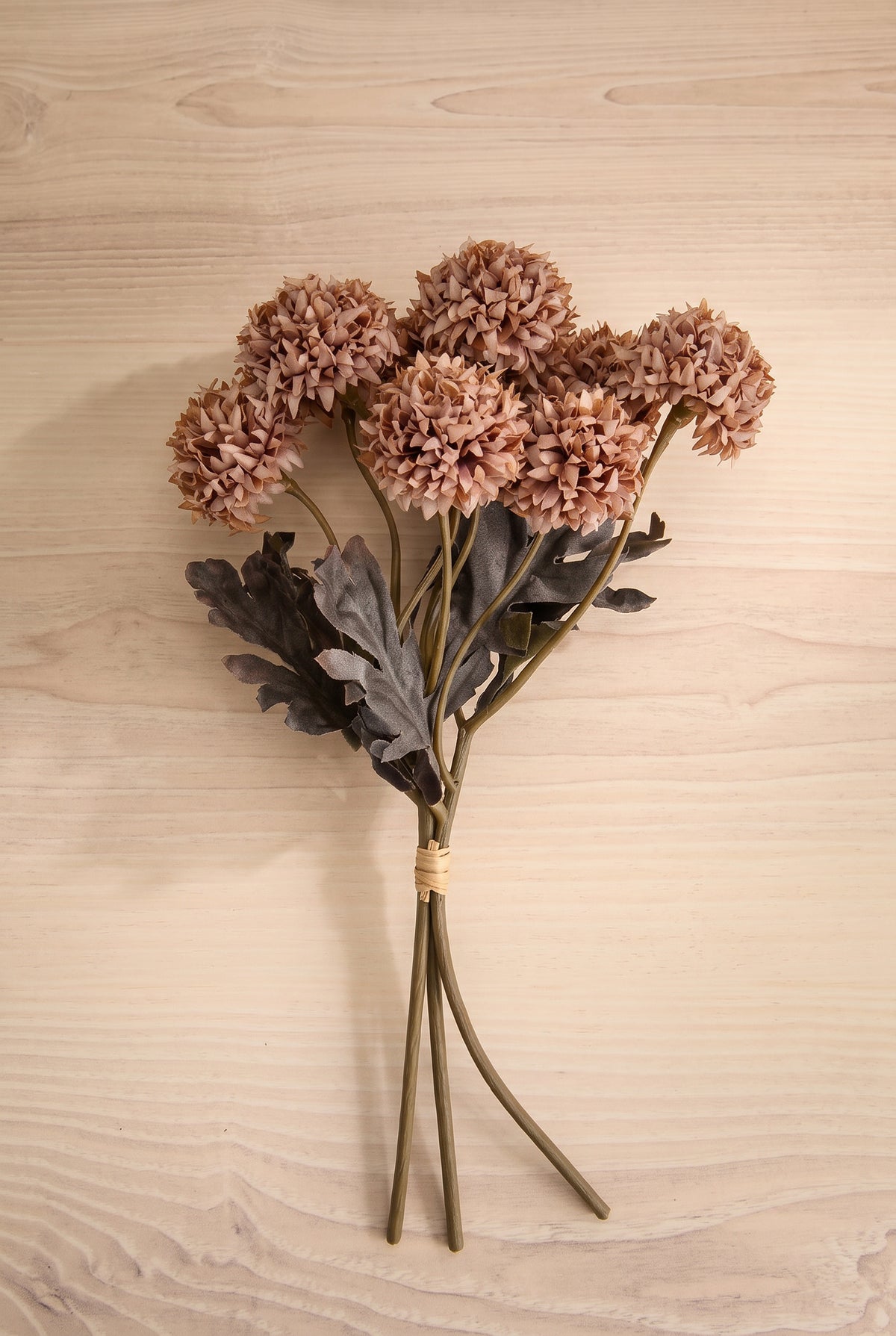 A bouquet of dried decorative artificial morandi flowers with dusty pink-brown round blooms, muted gray-green leaves, and long brown stems tied together with a beige band, placed on a light wood surface at la maison garçonne in montreal.
