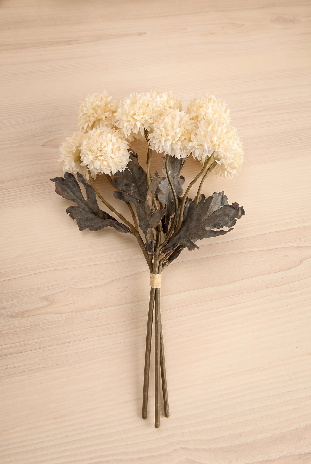 A bouquet of artificial cream-colored morandi-like flowers with dark gray leaves and brown stems, tied with a beige band and placed on a light wood surface at la maison garçonne in montreal.