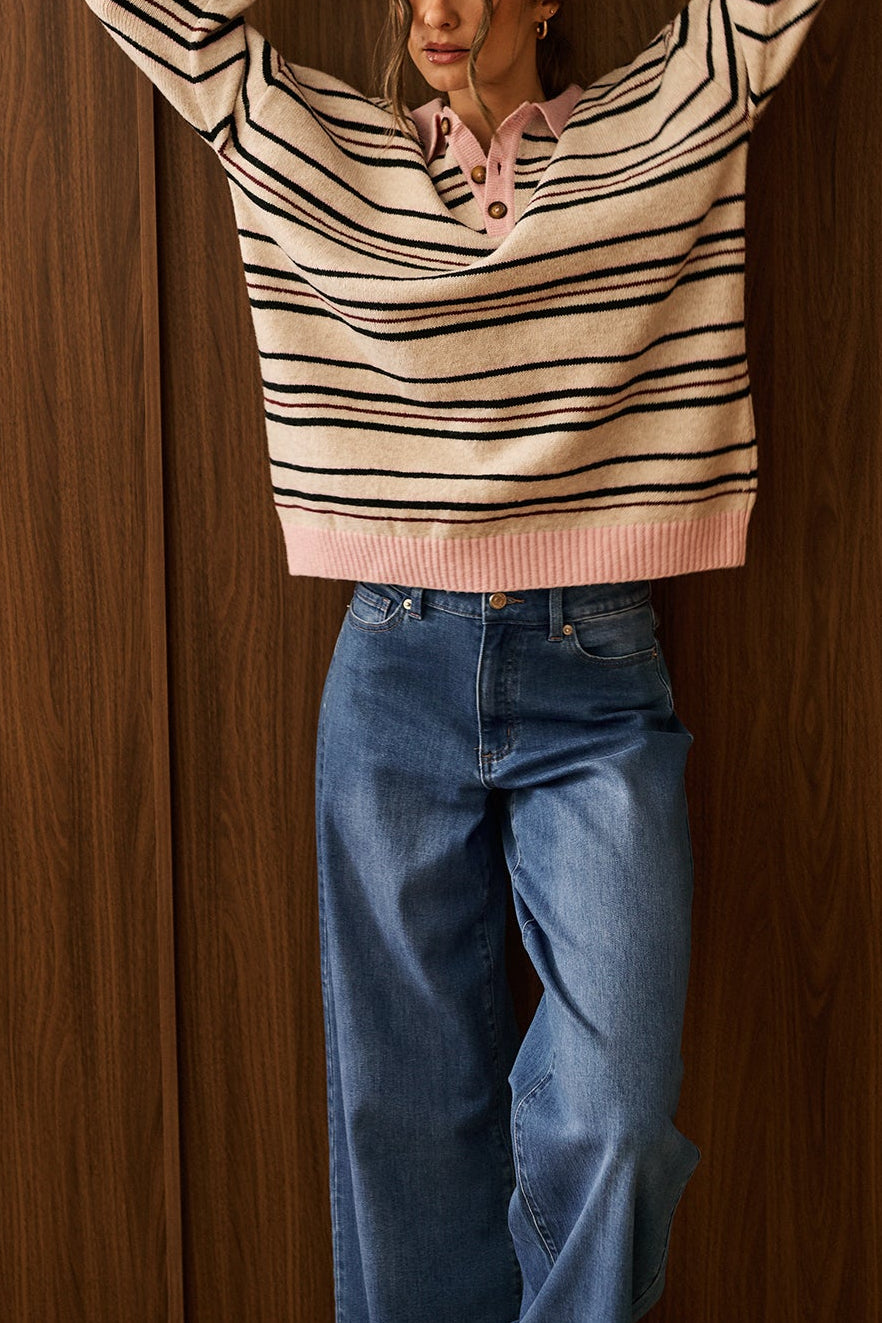 A woman stands against a dark wood-paneled wall wearing a beige knit sweater Gabbie with thin black horizontal stripes and a pink collar by dailystory Montreal
