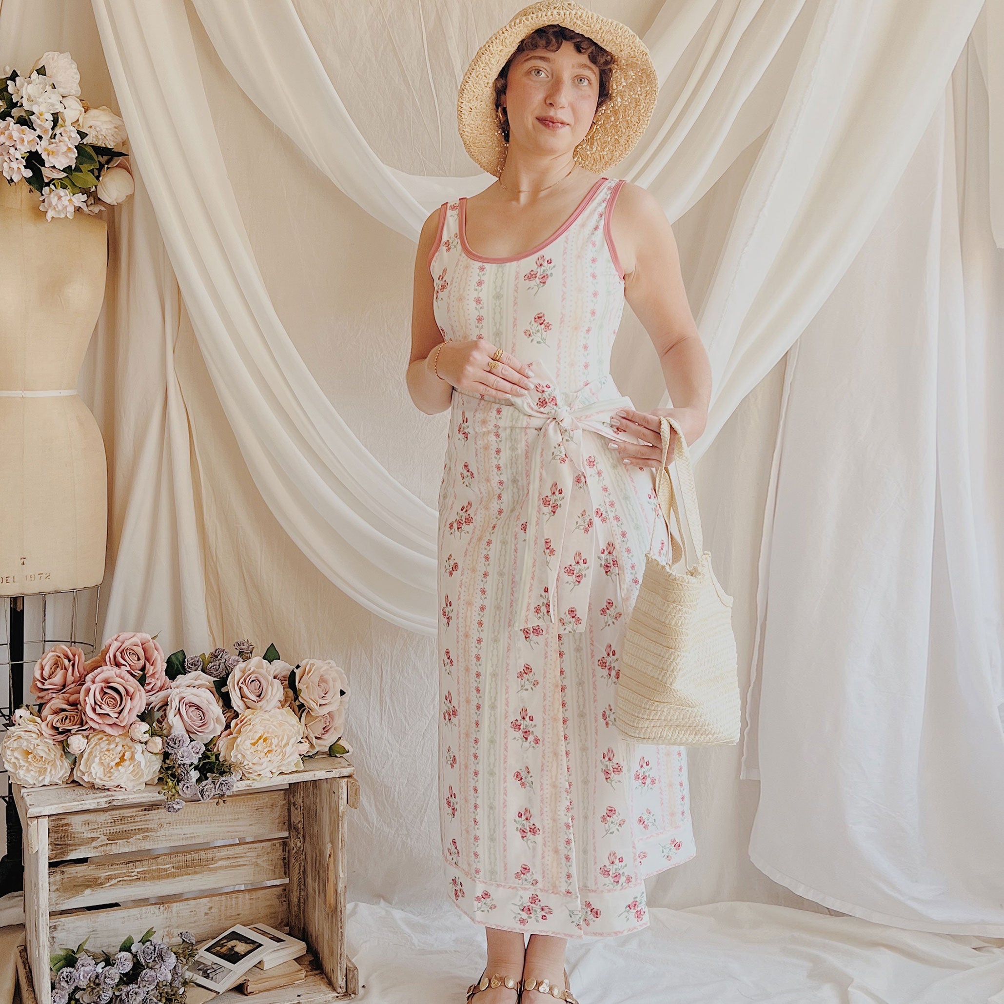 Woman in a floral dress and straw hat standing in front of white curtains with floral decorations.