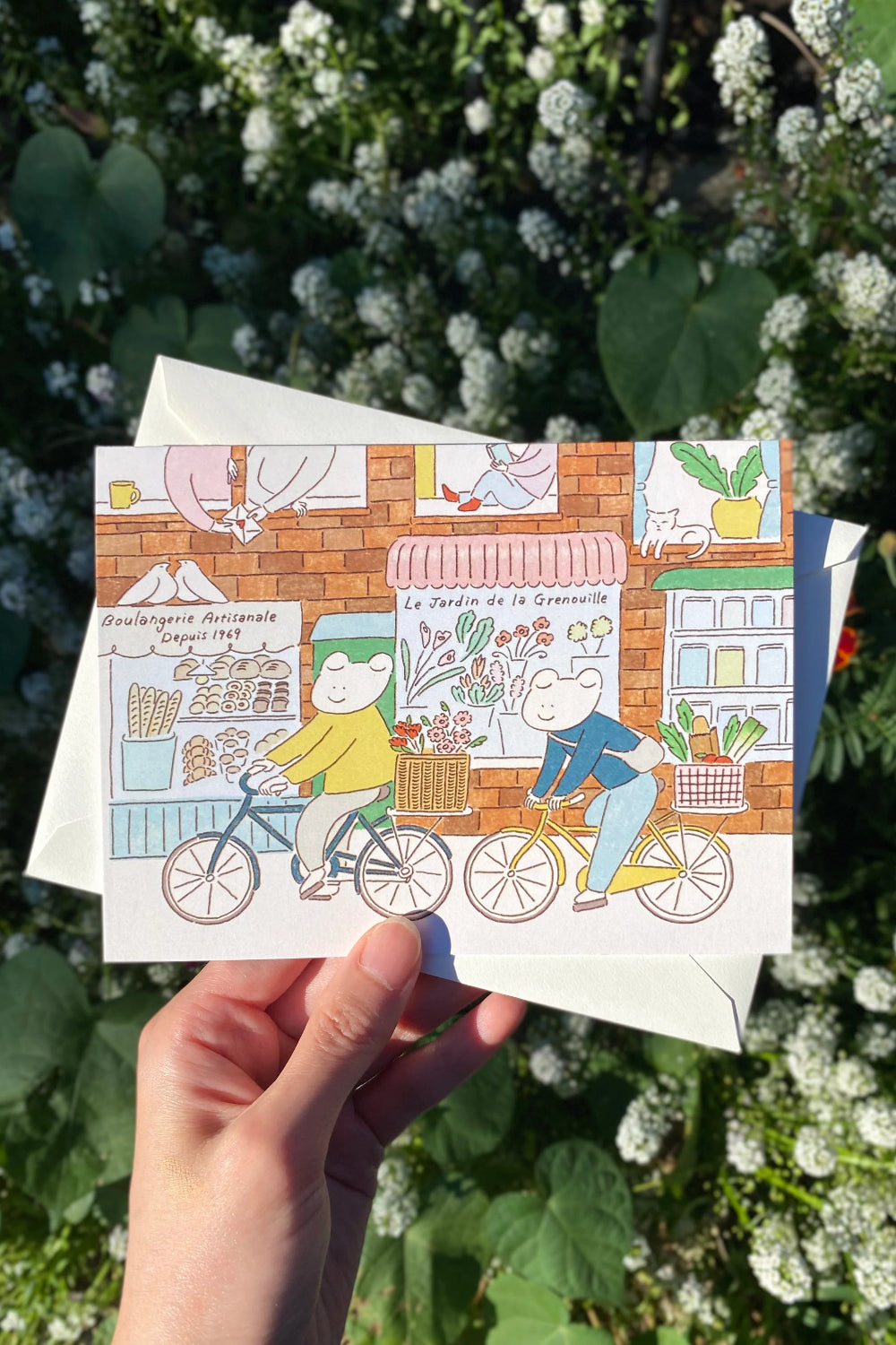 Illustrated postcard with two frogs on bicycles held by a hand in the city