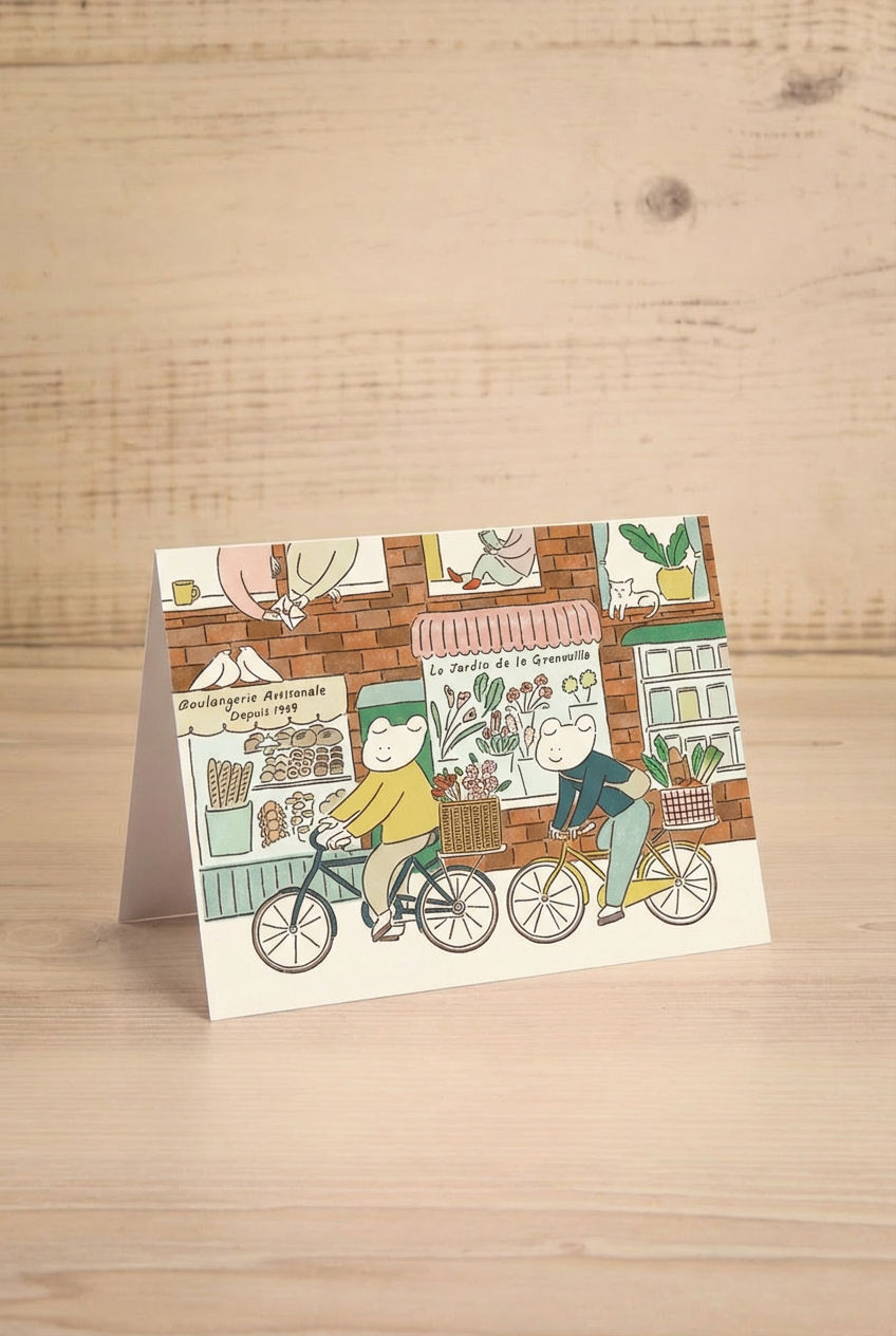 Illustrated greeting card with two grogs on bicycles in front of a store on a wooden surface