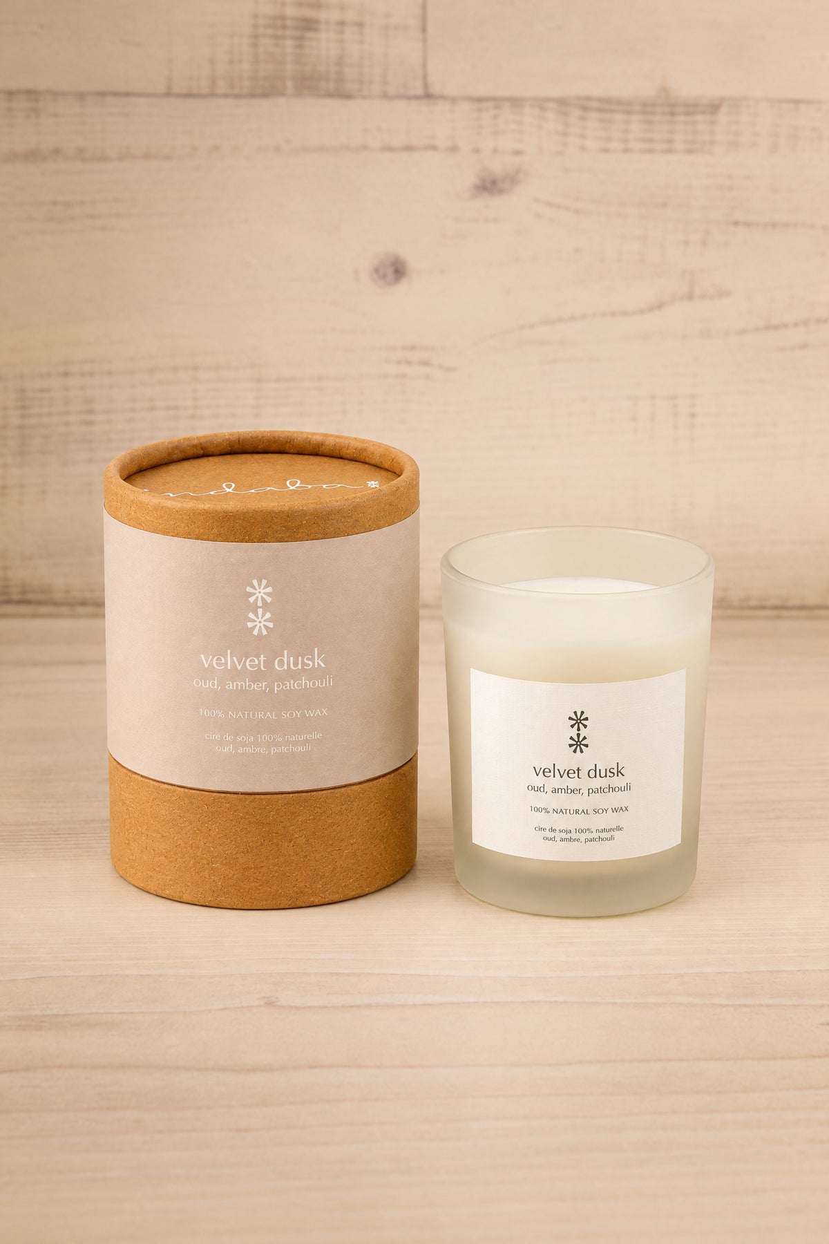 A jar candle and its kraft paper packaging box on a light wood background. The label reads 'velvet dusk' and 'pine and wood fragrance at la maison garçonne in montreal.