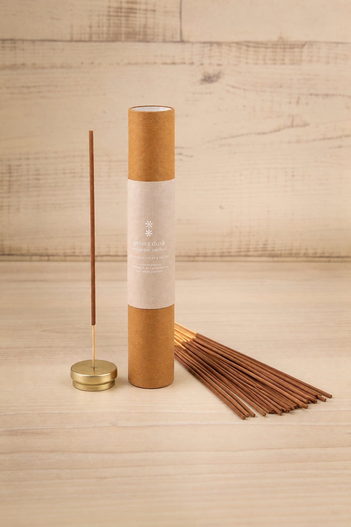 A tube of incense sticks with a kraft paper label stands next to a brass incense holder with a single stick lit, with several incense sticks fanned out on a light wood surface against a light wood plank background at la maison garçonne in montreal.