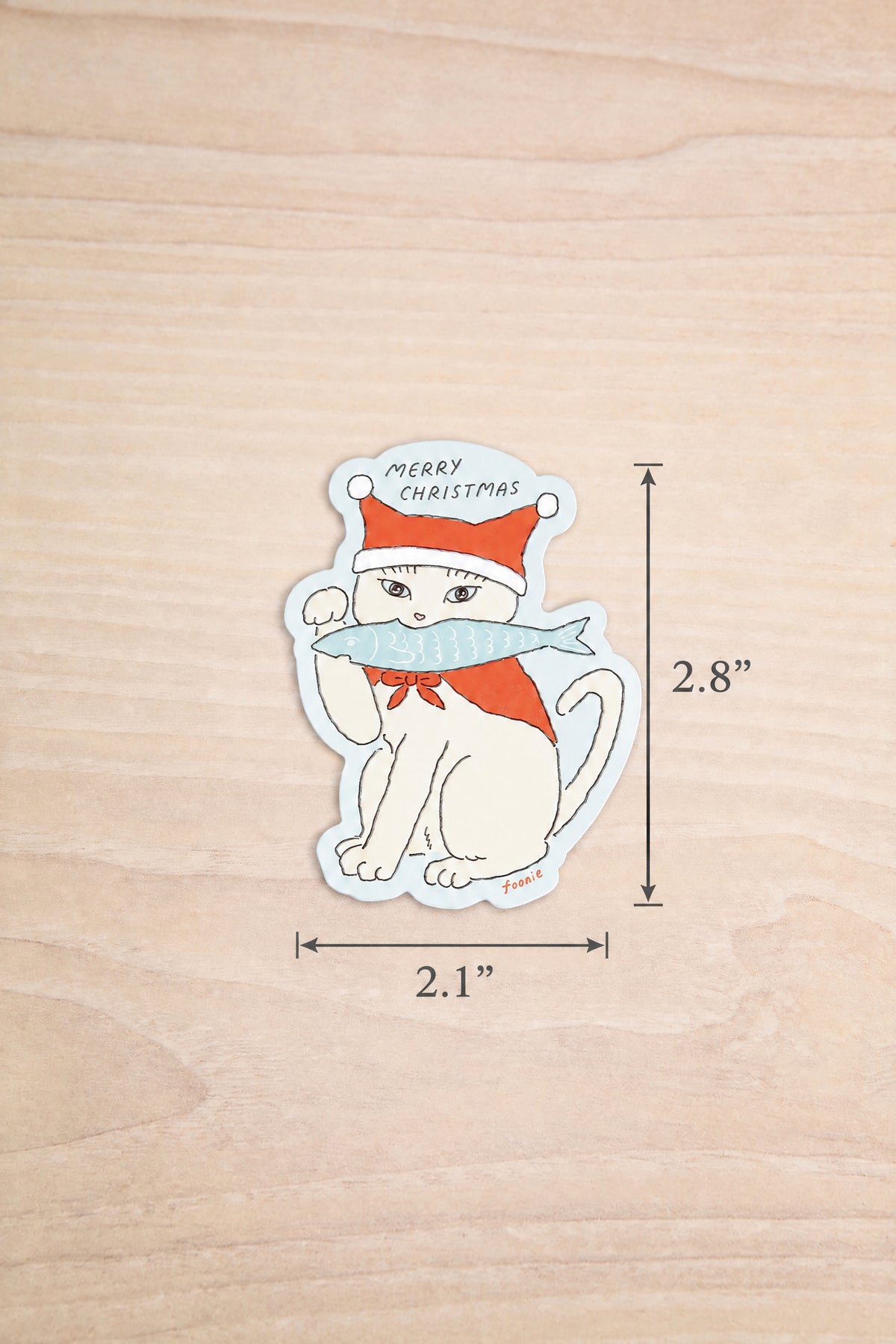 A die-cut sticker featuring a white cat wearing a red Santa hat and scarf from Foonie, with 'MERRY CHRISTMAS' text, shown on a light wood background with dimension markers of 2.1 inches by 2.