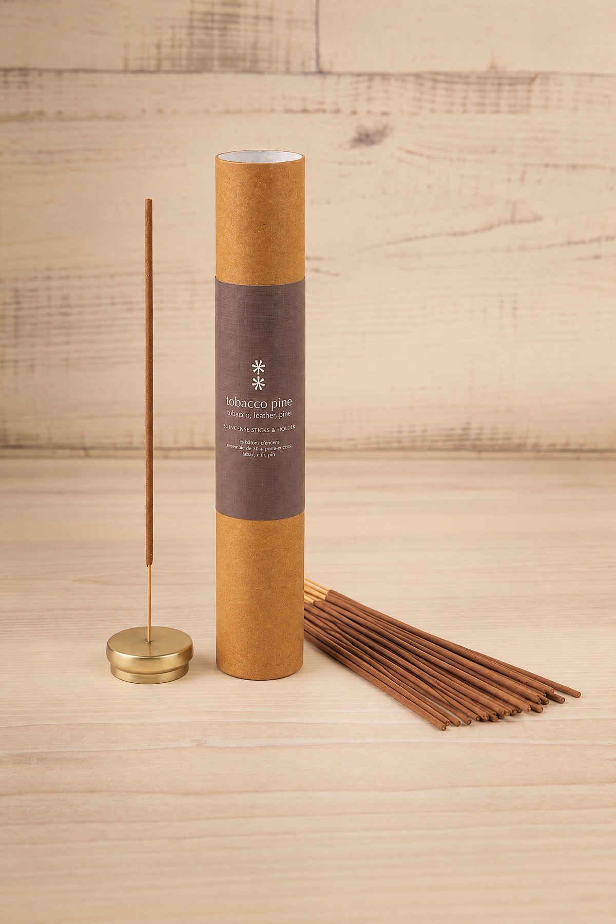 A set of incense sticks in a brown kraft paper tube with a dark brown label, shown next to a single incense stick on a brass holder and several loose incense sticks on a light wood background at la maison garçonne in montreal.