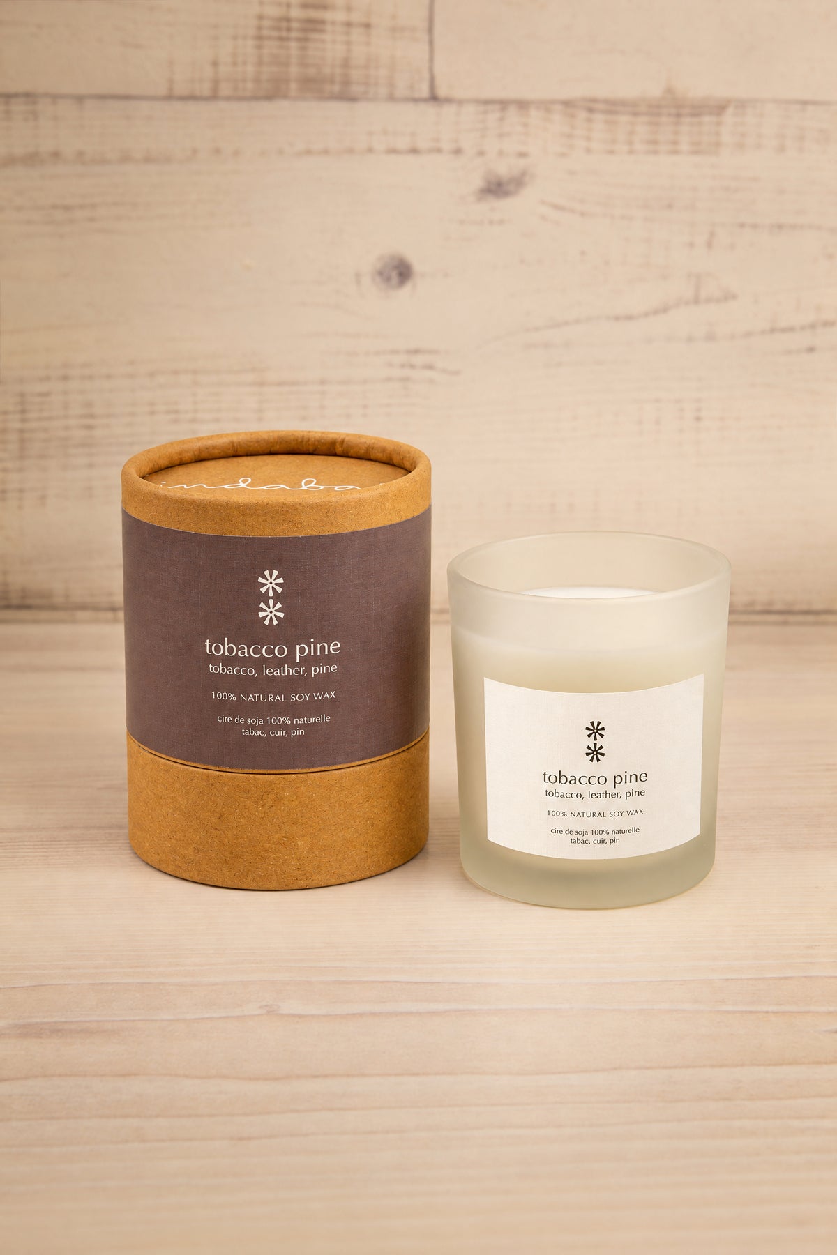 A tobacco pine scented candle shown next to its kraft paper and cardboard cylindrical packaging on a light wood plank surface at la maison garçonne in montreal.