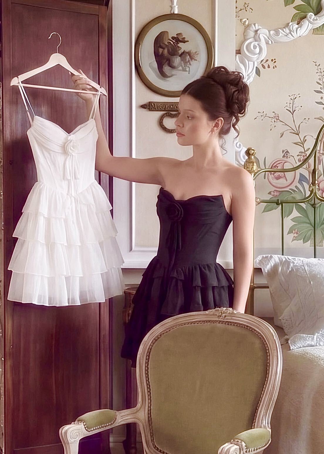 A woman in a black strapless dress stands in an ornate room holding a white tiered tulle dress on a hanger next to a wooden wardrobe at boutique 1861.