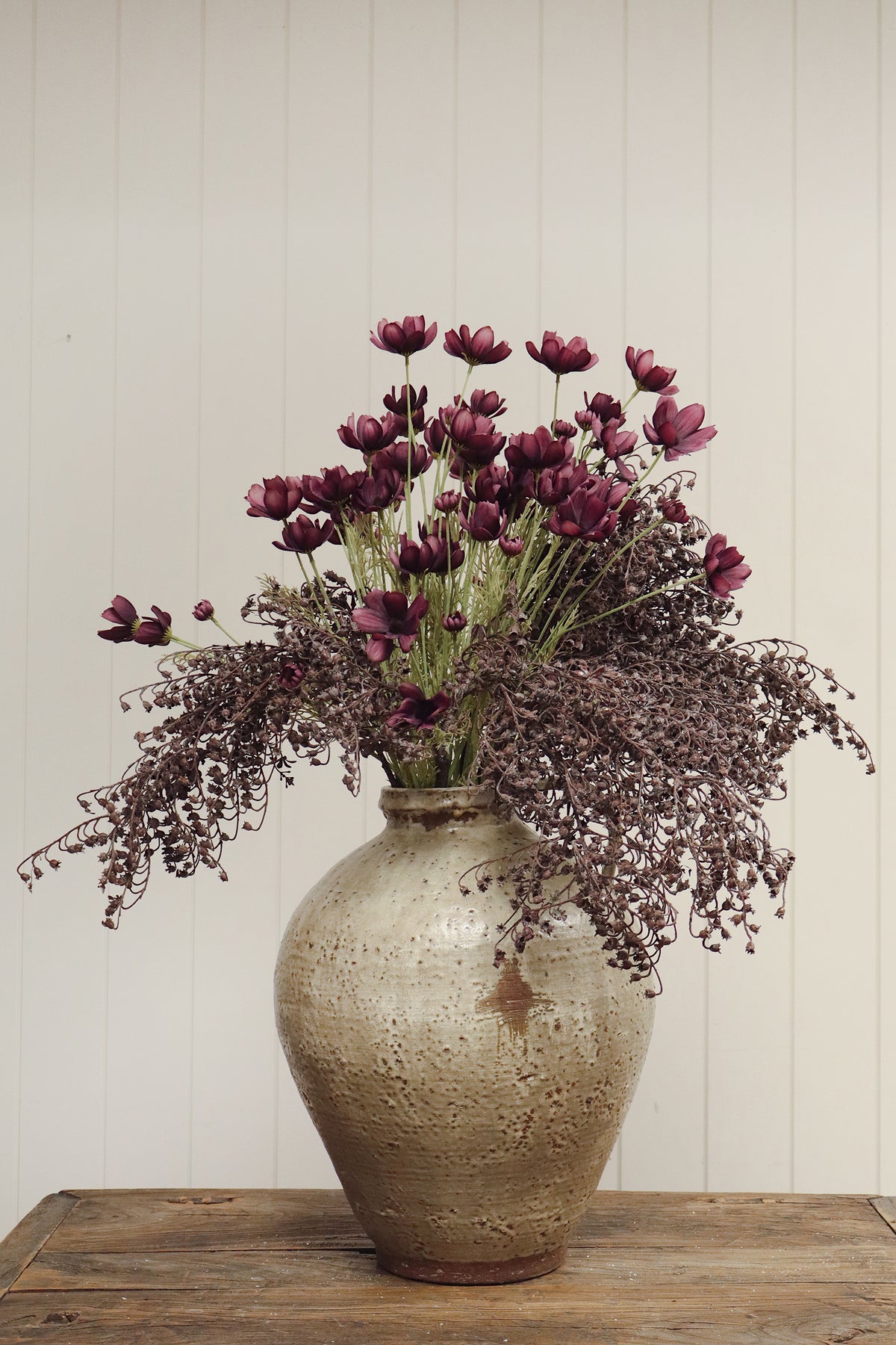 Vase with artificial deep purple calliopsis spray flowers with long green stems and feathery foliage, on a wood-grain surface at la maison garçonne in montreal.
