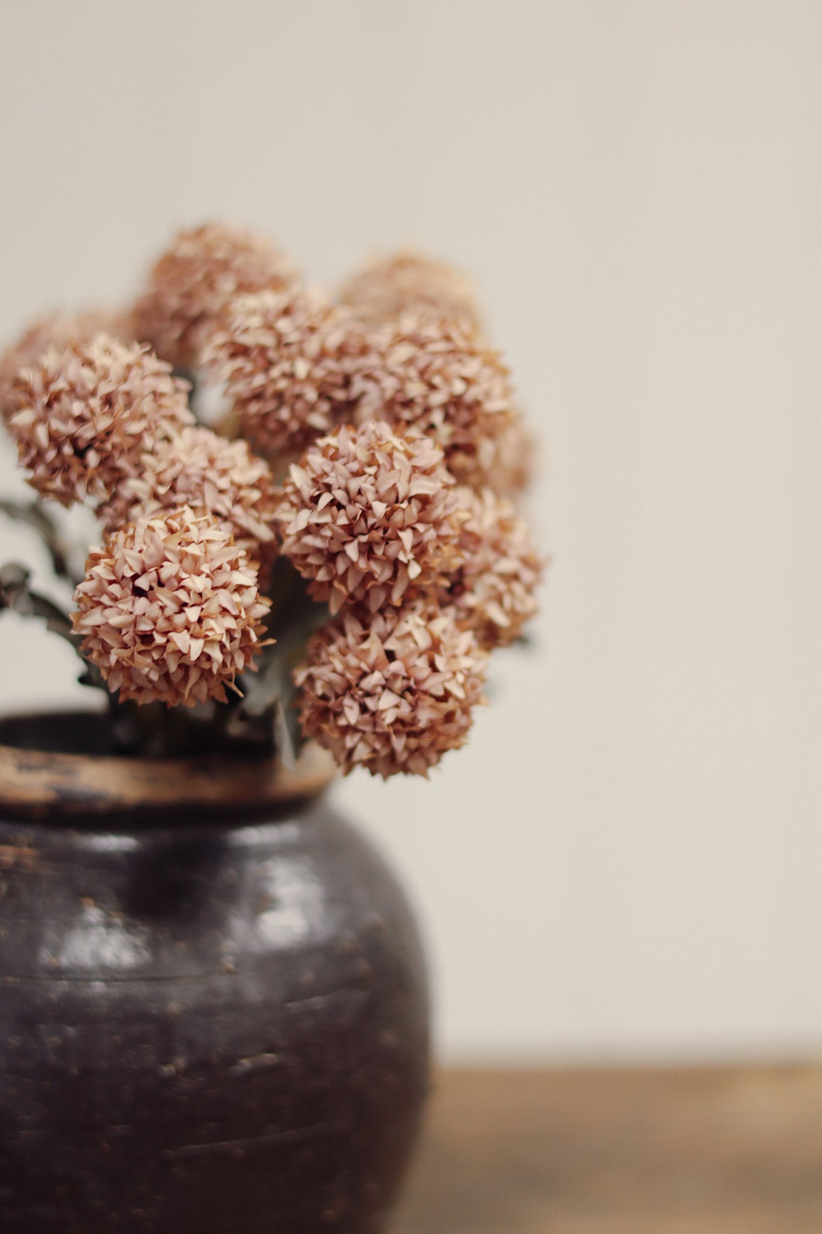 super close up on a decorative vase with decorative artificial morandi flowers with dusty pink-brown round blooms, muted gray-green leaves, and long brown stems tied together with a beige band, placed on a light wood surface at la maison garçonne in montreal.