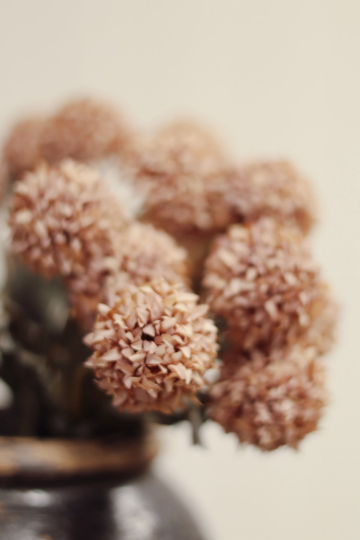 close up on a decorative vase with decorative artificial morandi flowers with dusty pink-brown round blooms, muted gray-green leaves, and long brown stems tied together with a beige band, placed on a light wood surface at la maison garçonne in montreal.