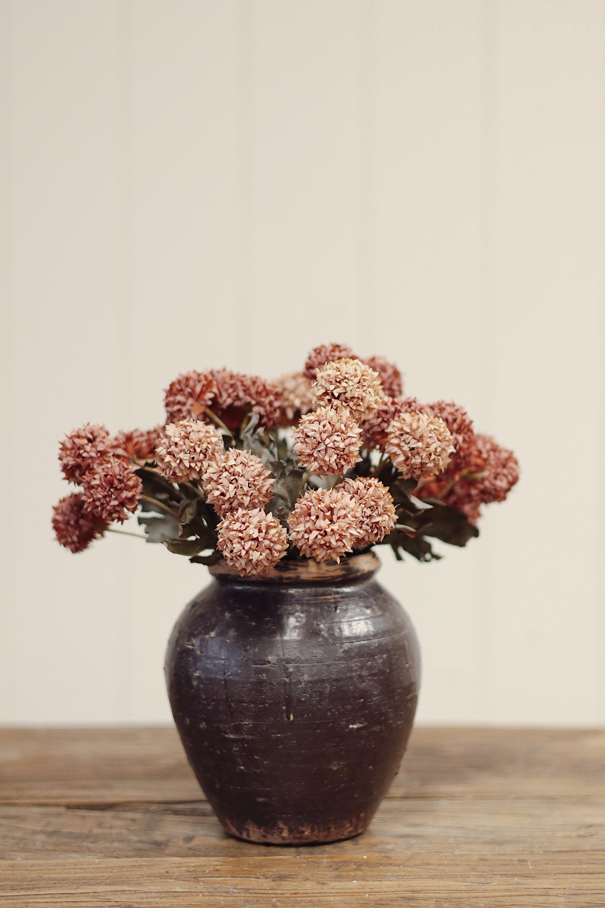 Decorative vase with decorative artificial morandi flowers with dusty pink-brown round blooms, muted gray-green leaves, and long brown stems tied together with a beige band, placed on a light wood surface at la maison garçonne in montreal.