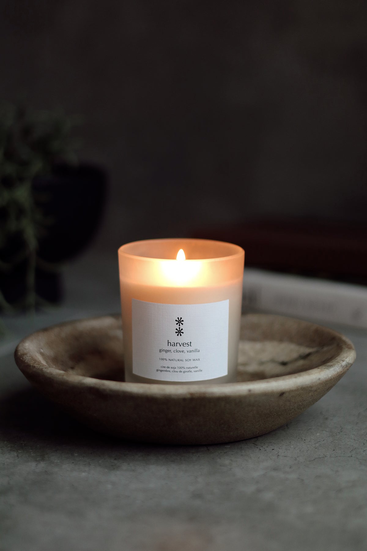 A lit candle in a white jar sits in a shallow rustic ceramic dish on a gray surface, with a dark blurred background at la maison garçonne in montreal. 