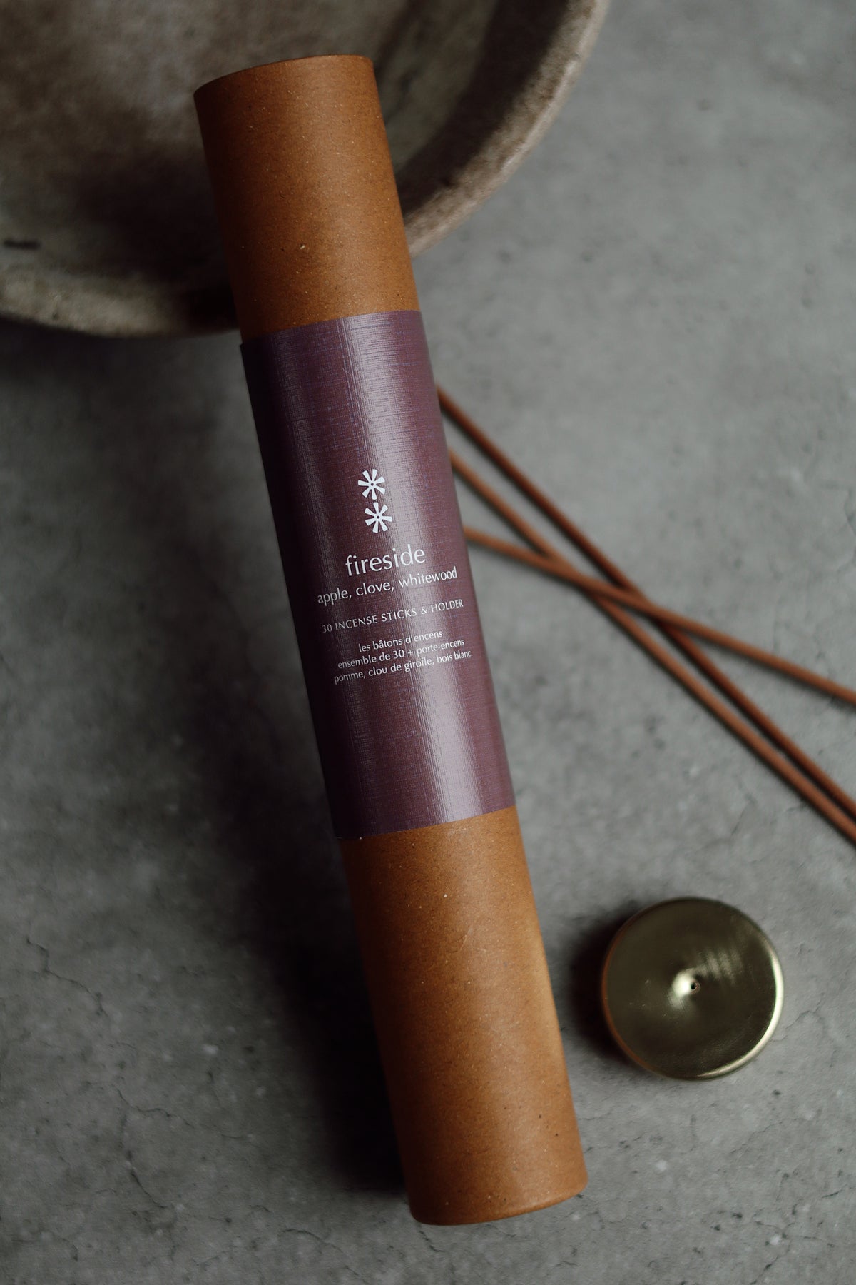 A brown kraft paper tube with a dark purple label reading 'fireside' and 'moss pine scented candle' lies on a gray stone surface, with a small brass lid and incense sticks nearby at la maison garçonne in montreal.