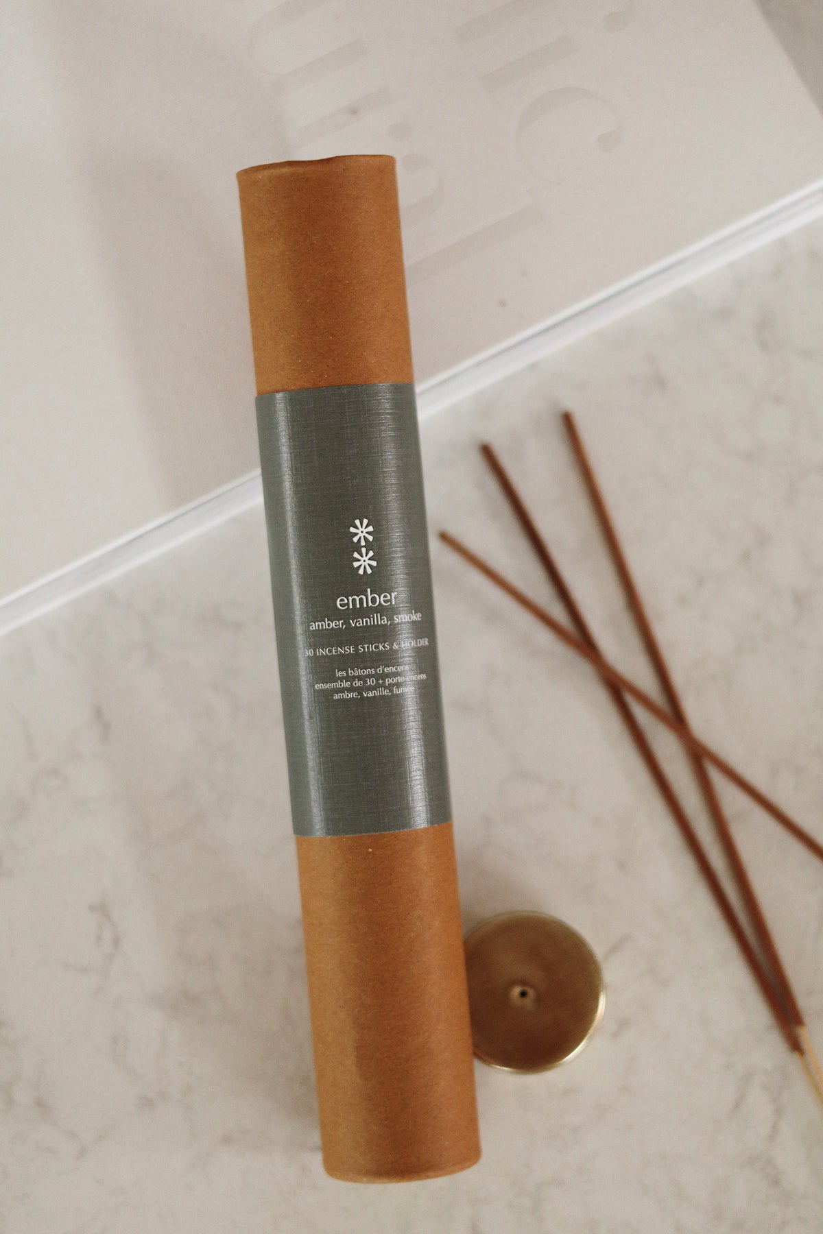 An incense stick set by the brand 'ember', featuring a brown kraft paper cylinder with a gray paper band label, shown next to loose incense sticks on a light marble surface at la maison garçonne in montreal.