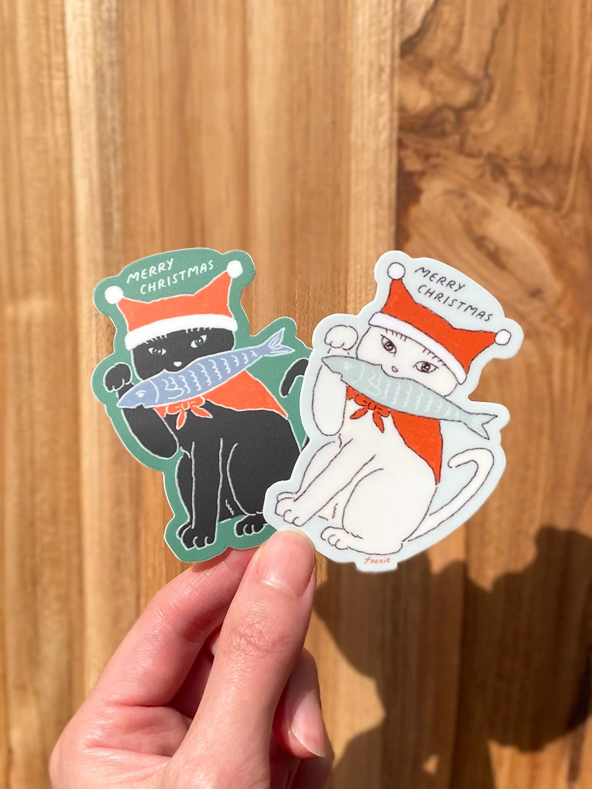 Two Christmas-themed die-cut stickers held by a hand against a wooden background. One sticker shows a black cat wearing a Santa hat and scarf while holding a fish, and the other shows a white cat wearing a Santa hat and scarf with a gray scarf.