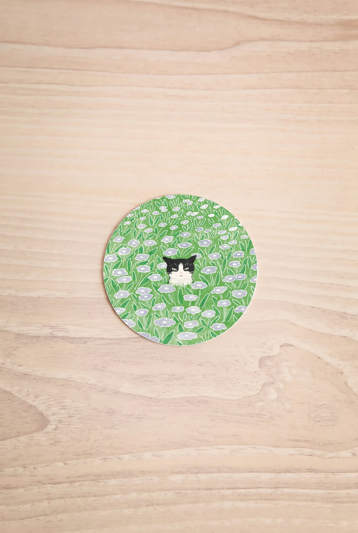 A round sticker featuring a green and white floral pattern with a small tuxedo cat in the center, illustrated by Foonie at la maison garçonne montreal