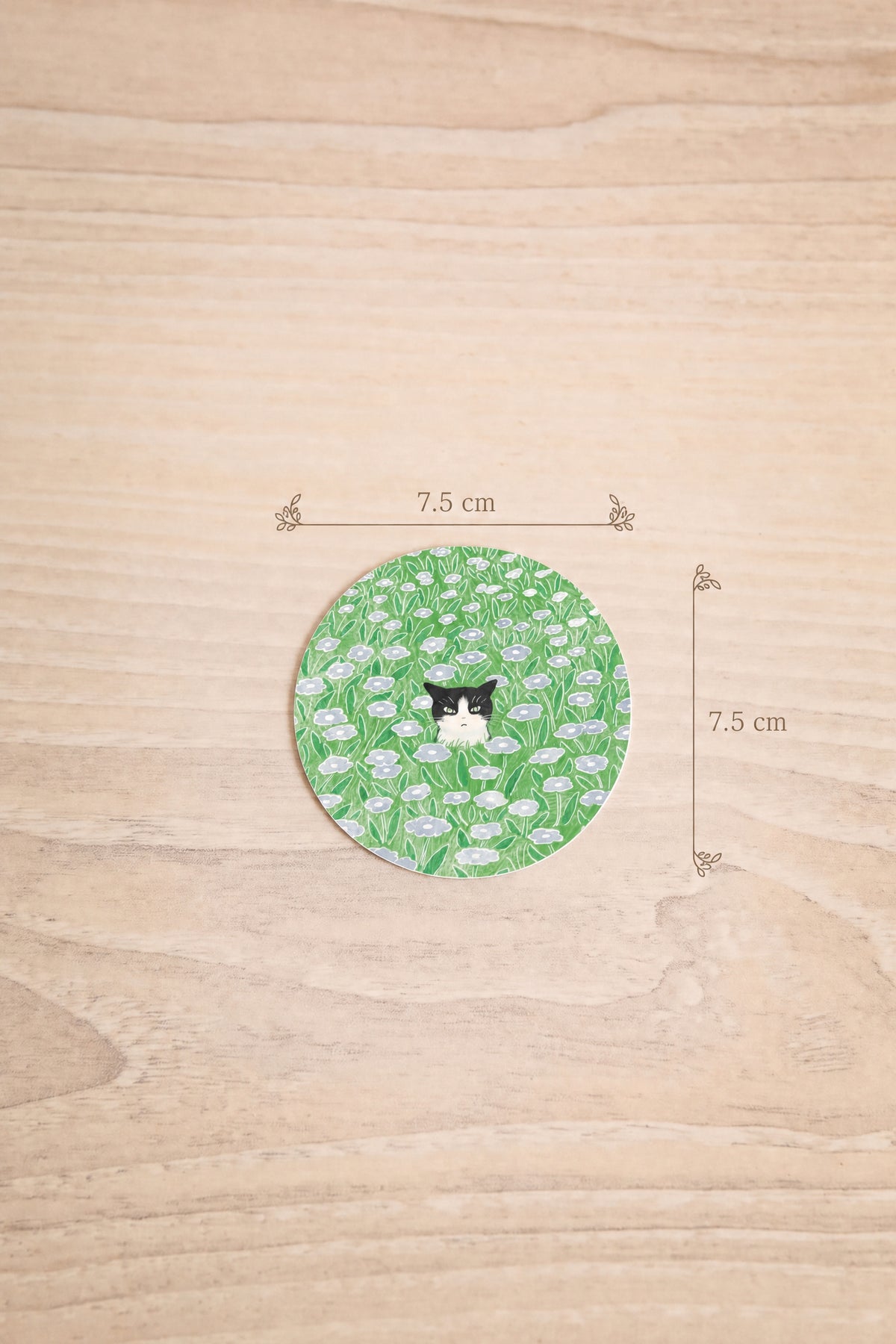 dimension 7.5cm x 7.5 cm of a round sticker featuring a green and white floral pattern with a small tuxedo cat in the center, illustrated by Foonie at la maison garçonne montreal