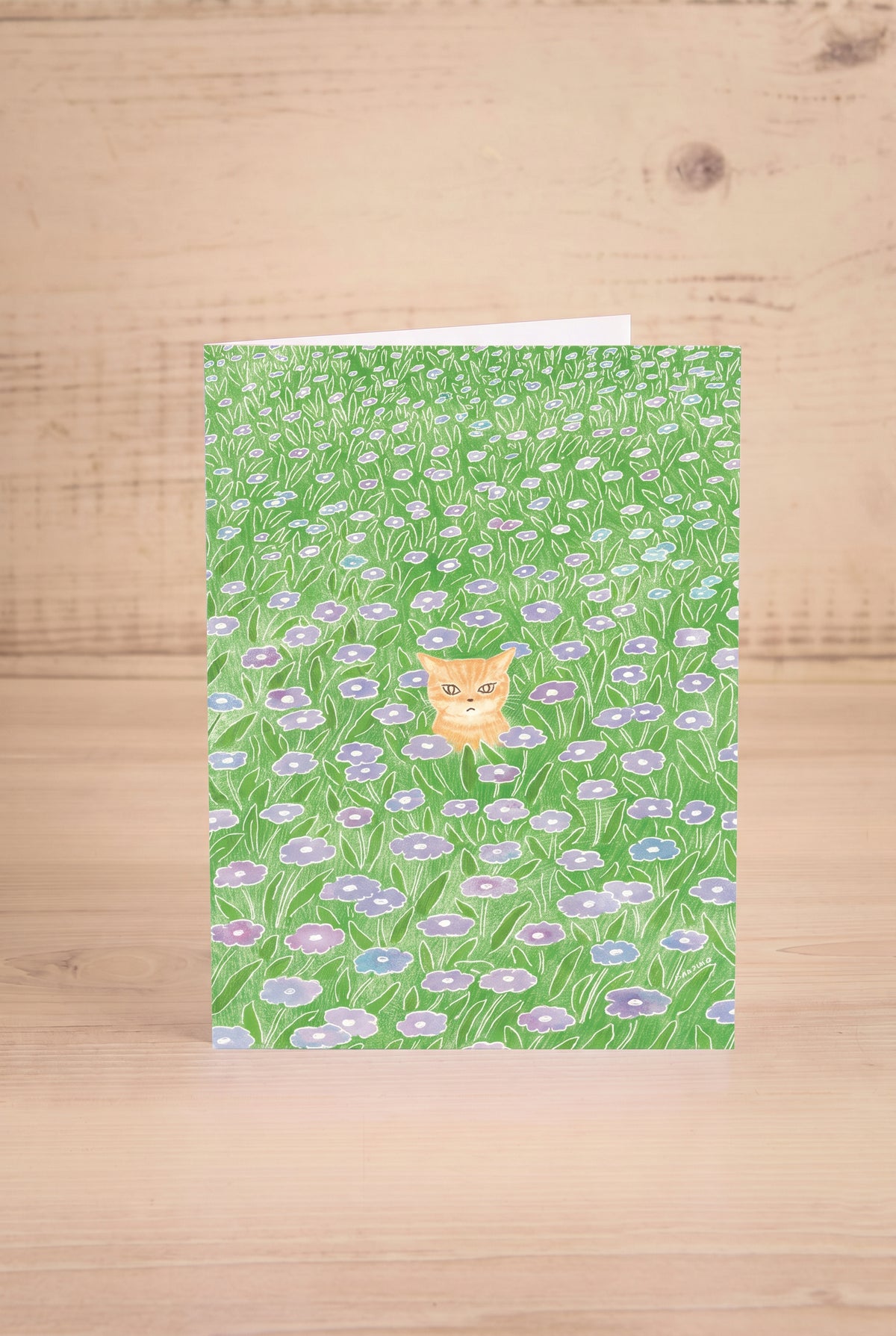 A vertical greeting card featuring a watercolor-style green field of flowers with a small orange cat by Foonie at maison garçonne in montreal